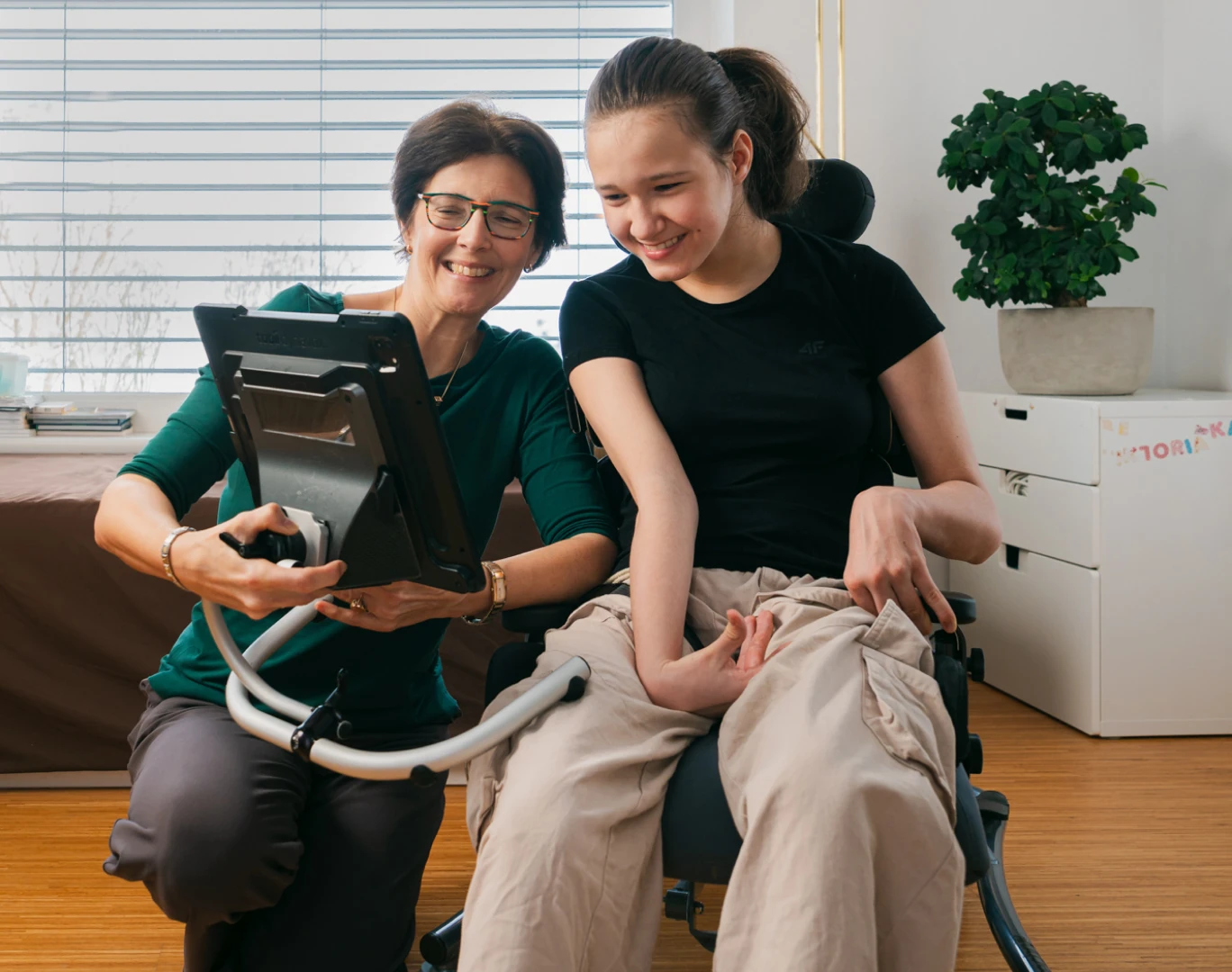 A mother and daughter are looking at a screen. It is attached directly to the daughter’s chair. Both are smiling.