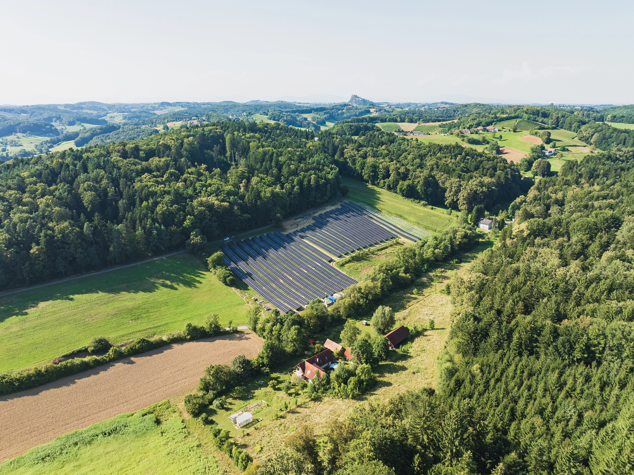 We look down on the photovoltaic system in Lödersdorf from above. It is nestled between forests. It is a beautiful day. The sun is shining.
