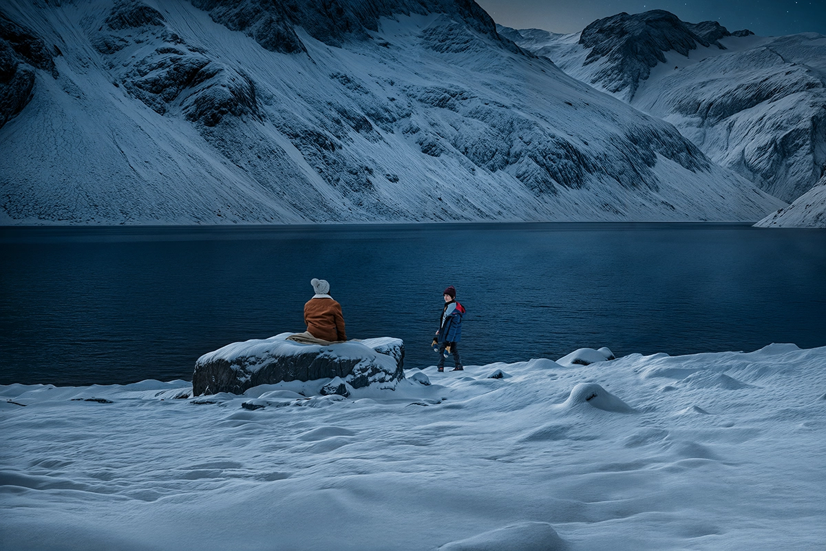 Mutter und Sohn spielen im Schnee an einem See. Beide tragen Mützen, Winterjacken und feste Schuhe. Die Sonne ist gerade untergegangen und es wird bereits Nacht.