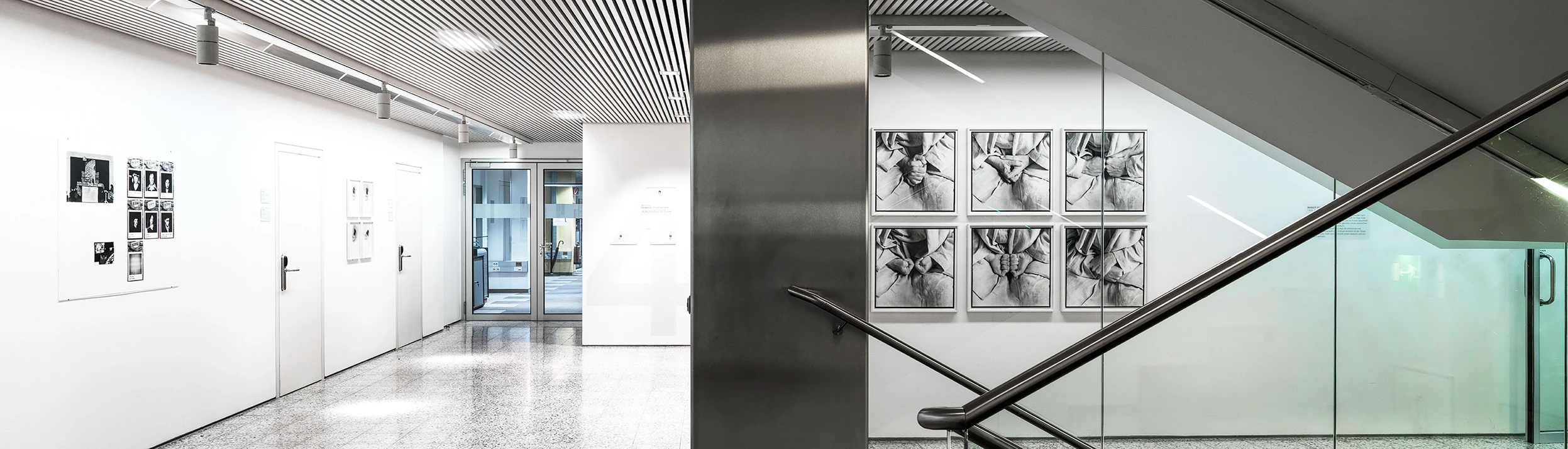 We look into the Vertikale Galerie in Vienna. We see a staircase decorated in light tones. Photographs are displayed on the walls.