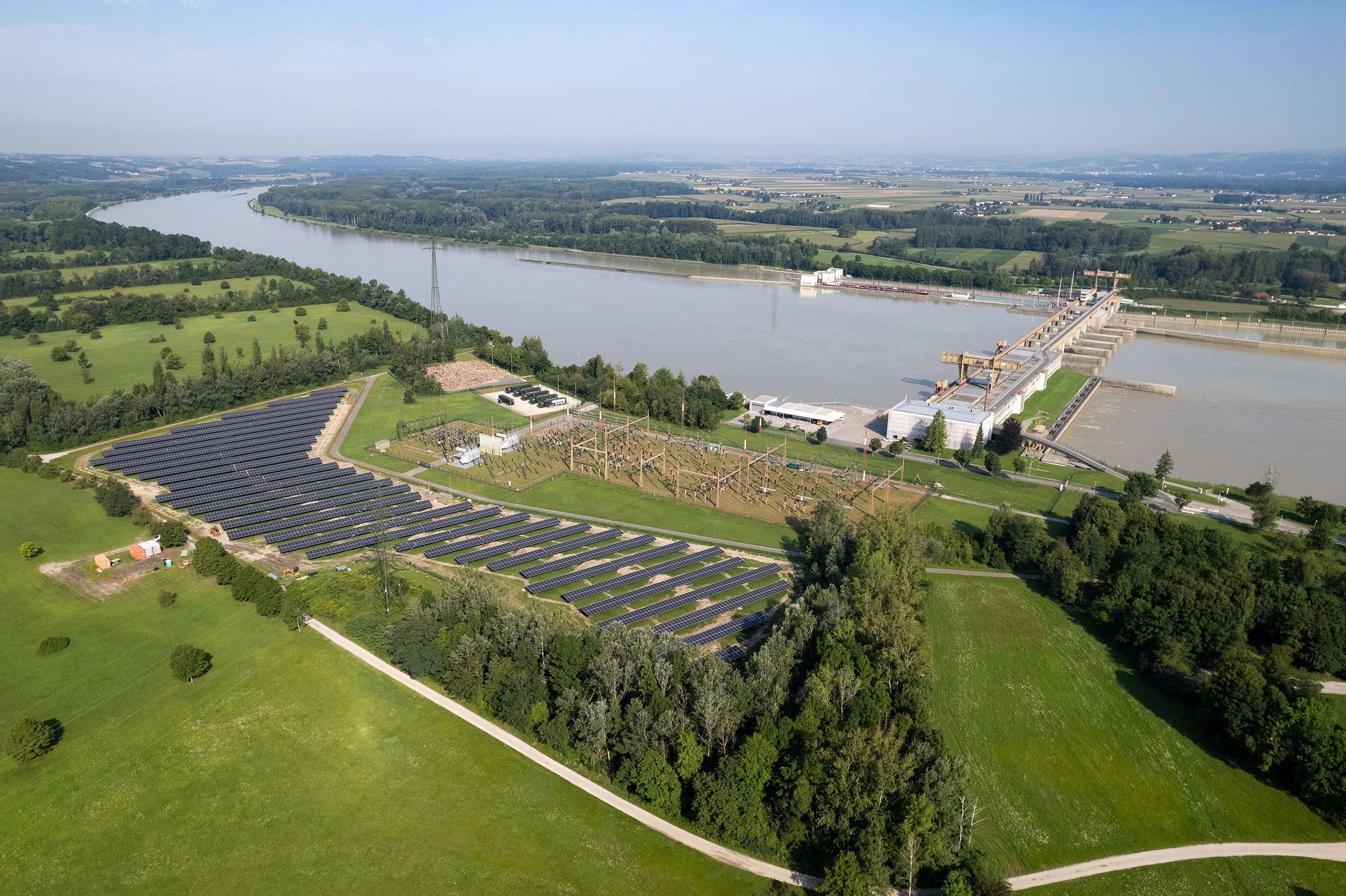 Das Kraftwerk Wallsee-Mitterkrichen von oben. Inklusive Batteriespeicher und Photovoltaikanlage. Die Umgeben ist grün und die Sonne scheint.