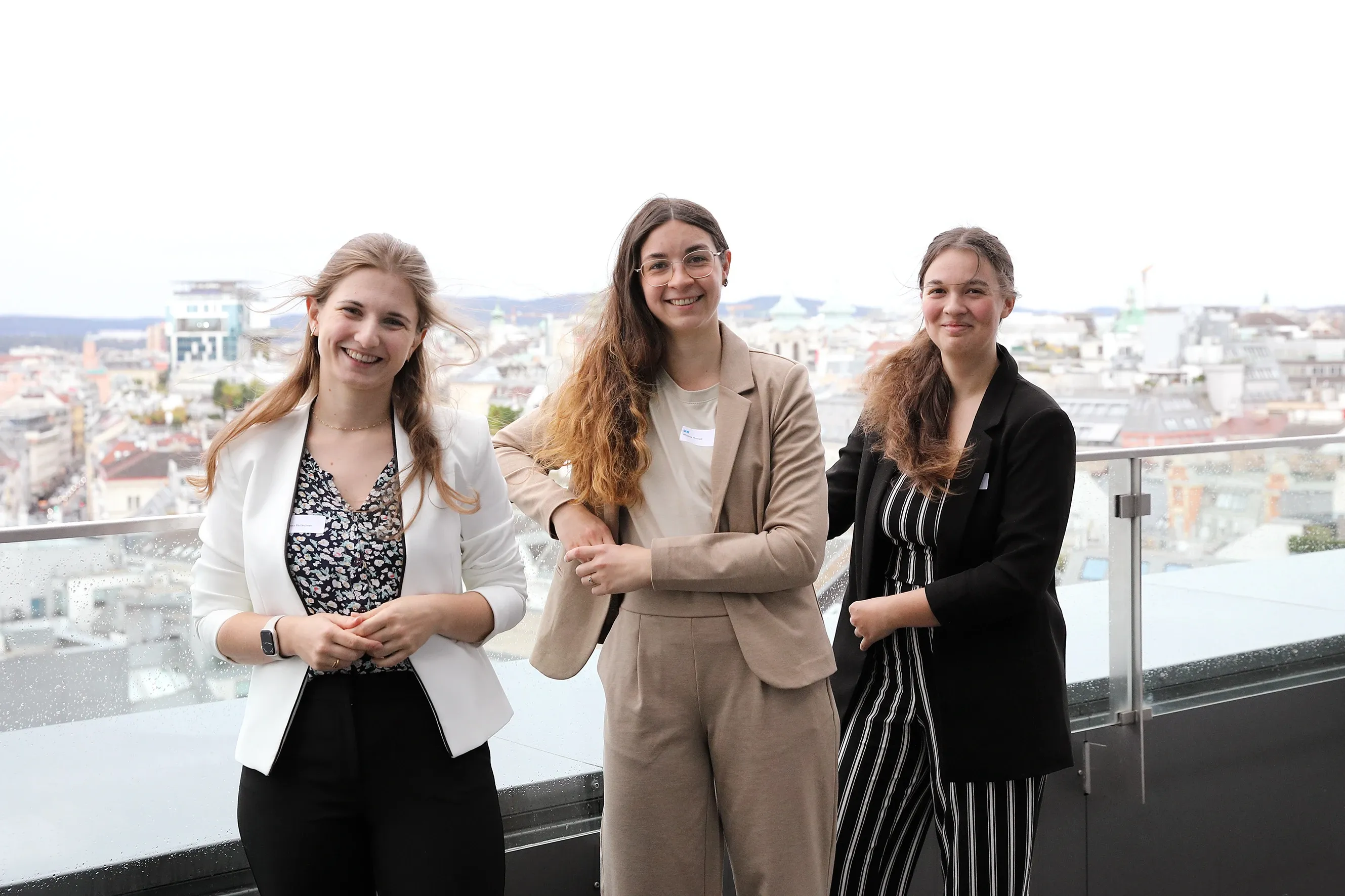 Drei junge Frauen lehnen an einer Balustrade aus Glas und lächeln in die Kamera. Die Frau links trägt ihr mittellanges dunkelblondes Haar offen und einen weißen Blazer, die Dame in der Mitte hat langes braunes Haar, lehnt ihren rechten Arm an die Balustrade und trägt einen beigen Anzug und die Dame ganz rechts trägt ihre langen Haar geschlossen, einen Nadelstreif Anzug und einen schwarzen Blazer.