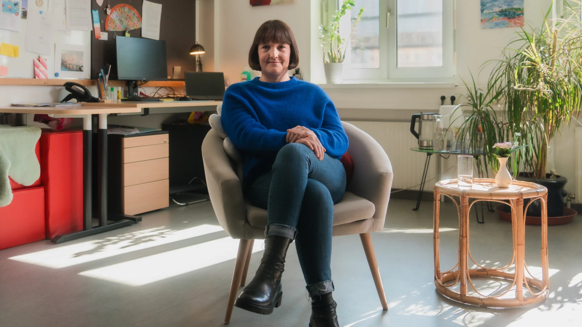 Eine Frau im blauem Pullover sitze auf einem Stuhl. Sie hat die Beine überschlagen und lächelt in die Kamera.