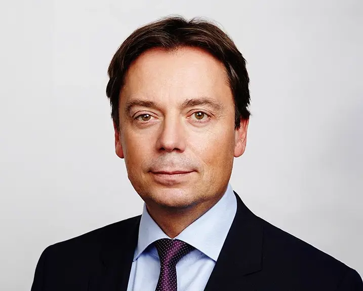 Andreas Wollein has dark brown hair and brown eyes. He is wearing a light blue shirt, a purple-coloured tie with small white dots and a black suit.