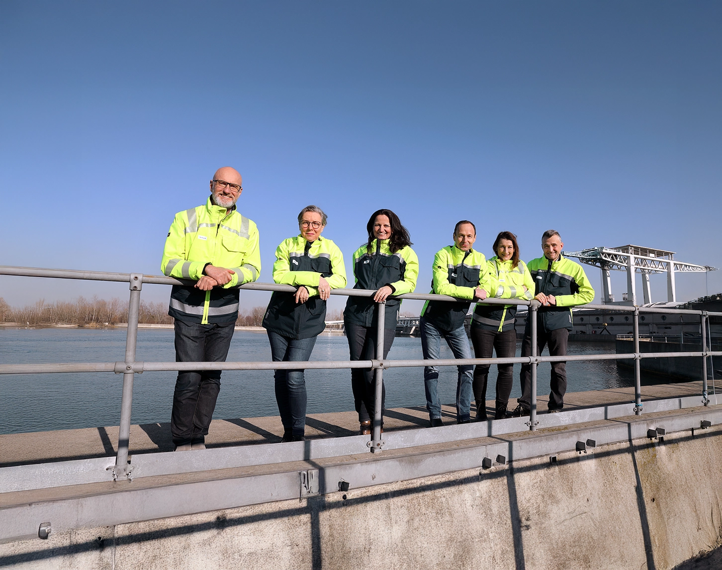 The entire VERBUND press team stands in front of a hydropower plant wearing protective clothing and smiling into the camera in the sunshine. The individual press officers are wearing protective clothing in bright yellow and dark blue with reflective stripes. They are leaning against a guardrail.