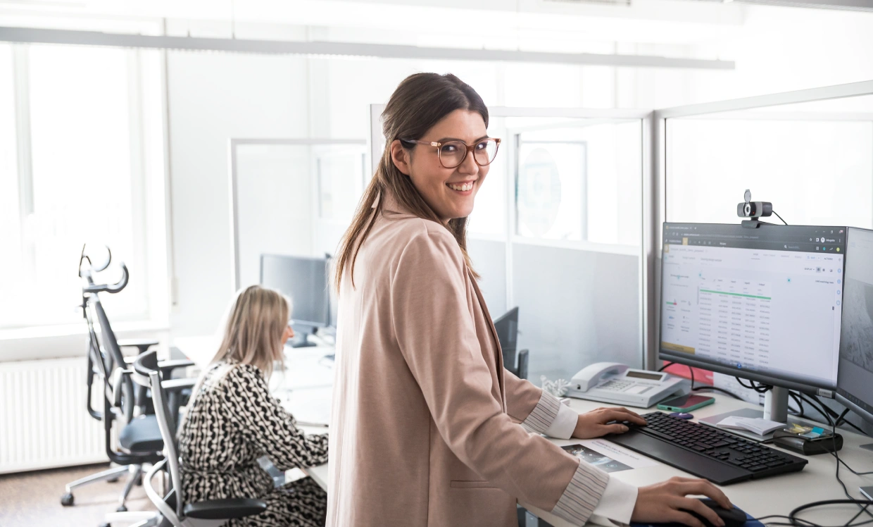 Person im roséfarbenen Blazer steht an einem Schreibtisch im Großraumbüro und arbeitet an einem Computer mit großem Bildschirm; im Hintergrund sitzt eine weitere Person im gemusterten Kleid an einem Arbeitsplatz, umgeben von Bürostühlen, Monitoren und hellem Tageslicht durch große Fenster.