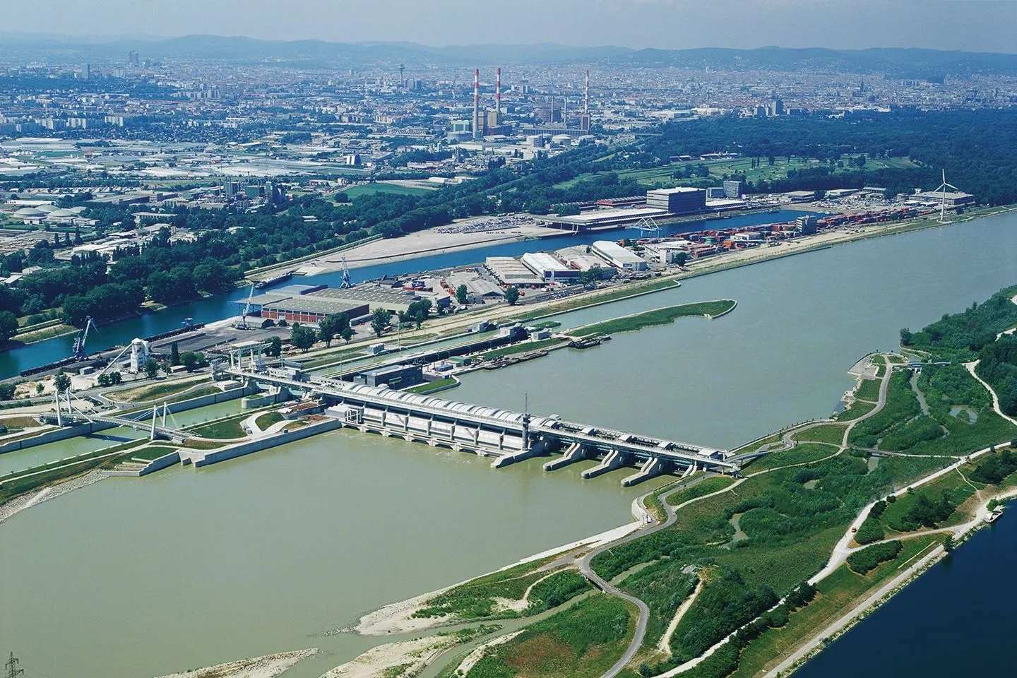 Zu sehen ist eine historische Aufnahme des Kraftwerk Freudenau an der Donau in Wien aus der Vogelperspektive. Im Vordergrund die Donau und das Kraftwerk, dahinter ist die Stadt Wien zu sehen. Ein sonniger Himmel ohne Wolken darüber.