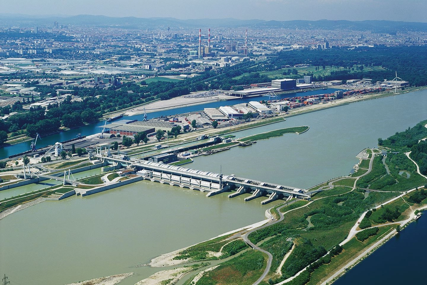 This is a historical photograph of the Freudenau power station on the Danube in Vienna from a bird's eye view. The Danube and the power station can be seen in the foreground, with the city of Vienna in the background. A sunny sky without clouds above.