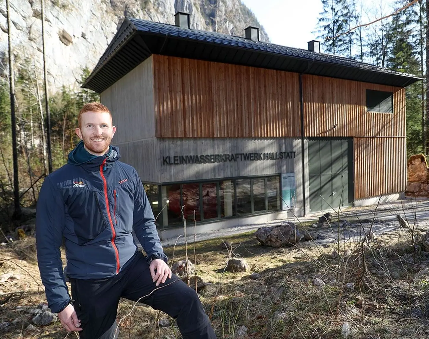 Ein Mann steht lächelnd vor einem Gebäude mit einer Holzfassade, auf dem "Kleinwasserkraftwerk Hallstatt" steht.