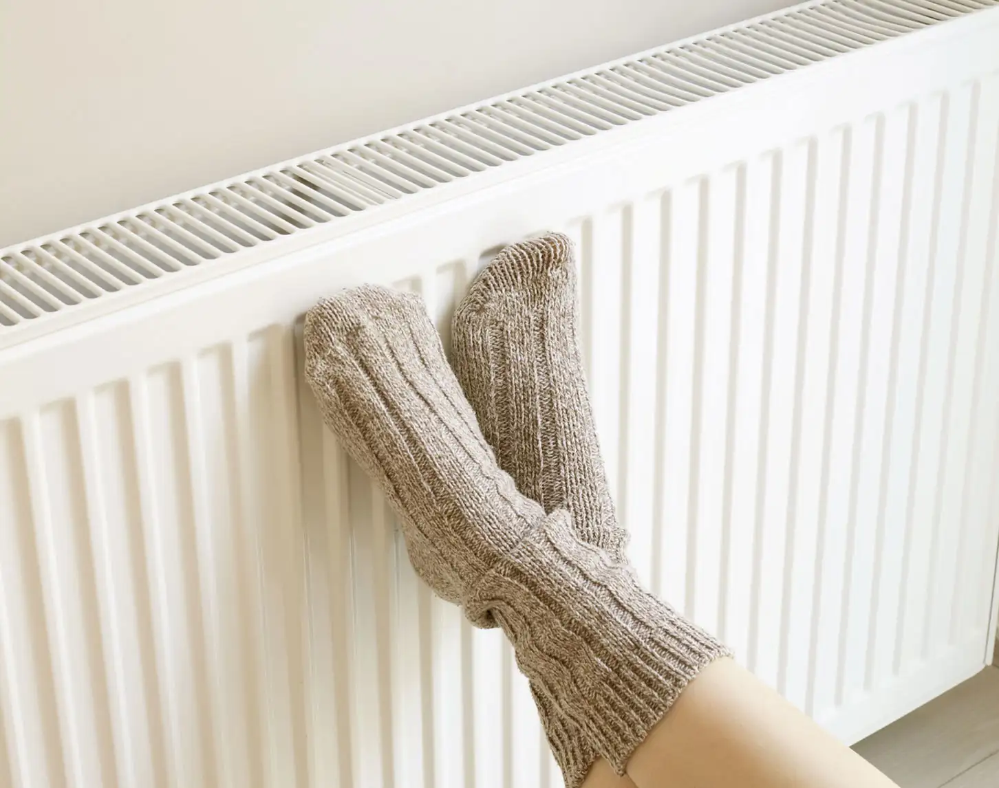 Überkreuzte Füße in dicken Stricksocken, die an einen Heizkörper angelehnt sind. Ein weißer Störer "5 Monate Gratis-Gas bei mind. 1 Jahr Laufzeit" und ein blauer Störer "Jetzt wechseln"