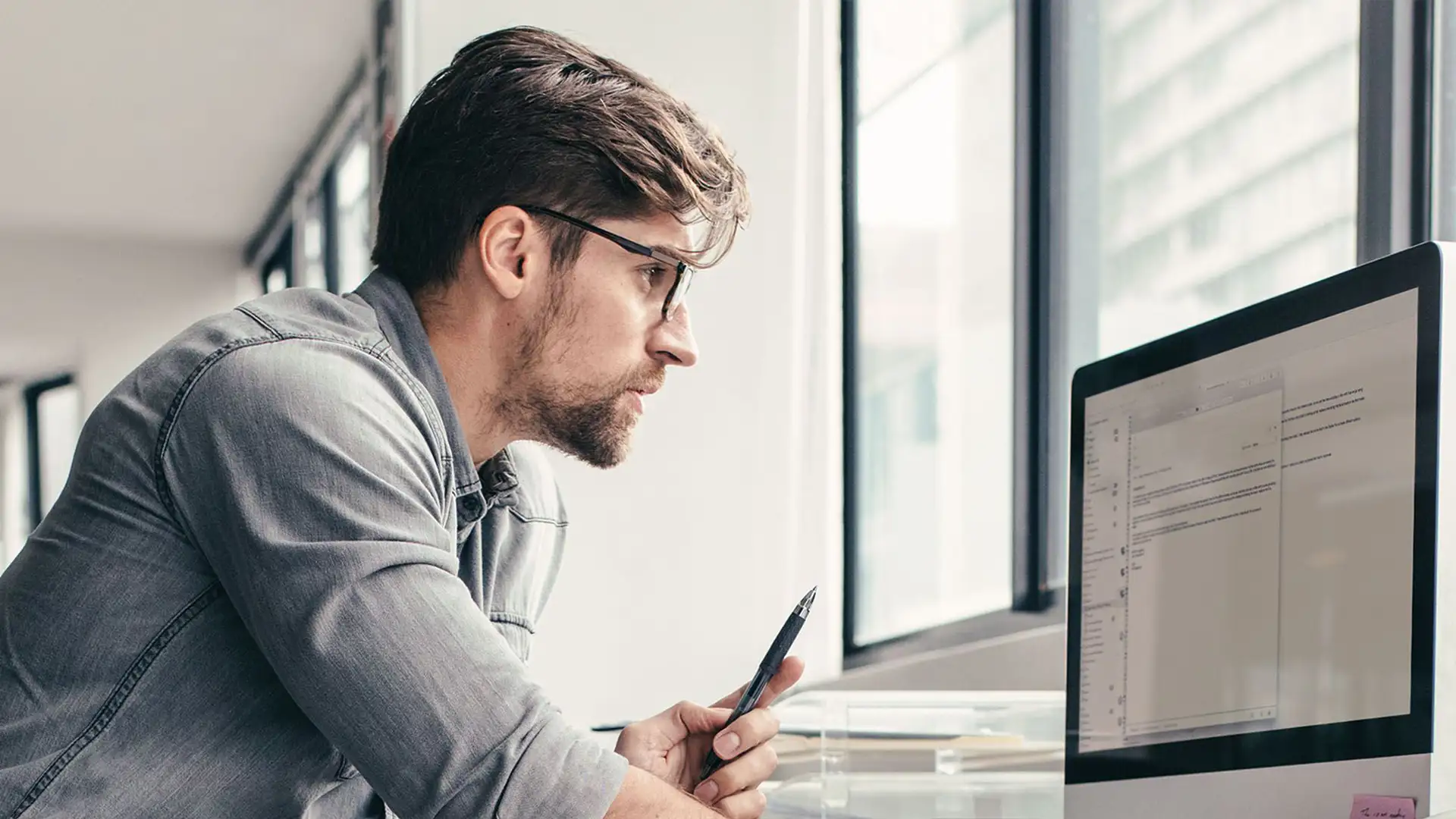 Businessman working attentively at a computer in a modern office. He holds a pen in his hand and is checking information important for optimizing Verbund business customers and their electricity tariffs.
