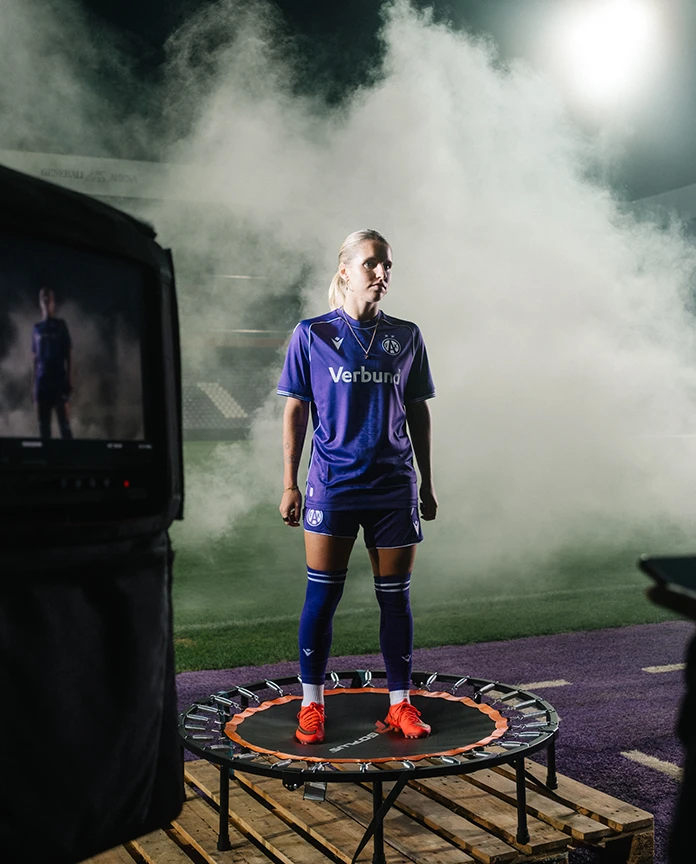 Eine Fußballspielerin im violetten VERBUND-Trikot steht auf einem Trampolin im Stadion, umgeben von Rauch.