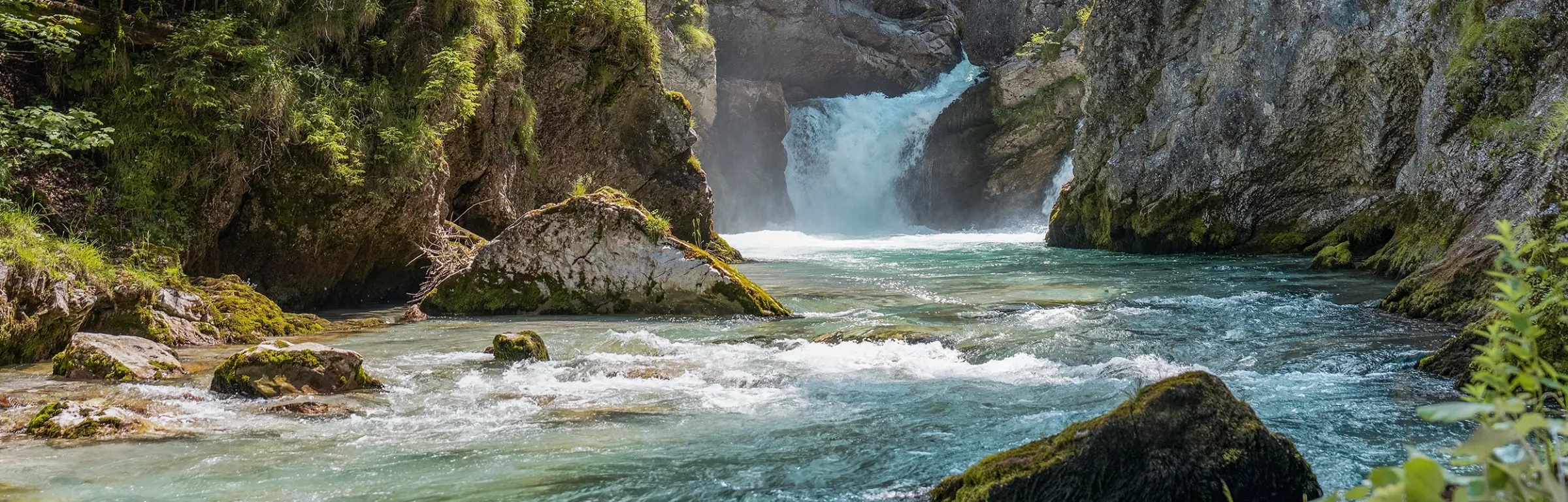 Man sieht einen rauschenden Bergfluss der sich seinen Weg durch die Felsen sucht. Die Sonne scheint auf karge Felsen, die mit Moos überzogen sind.
