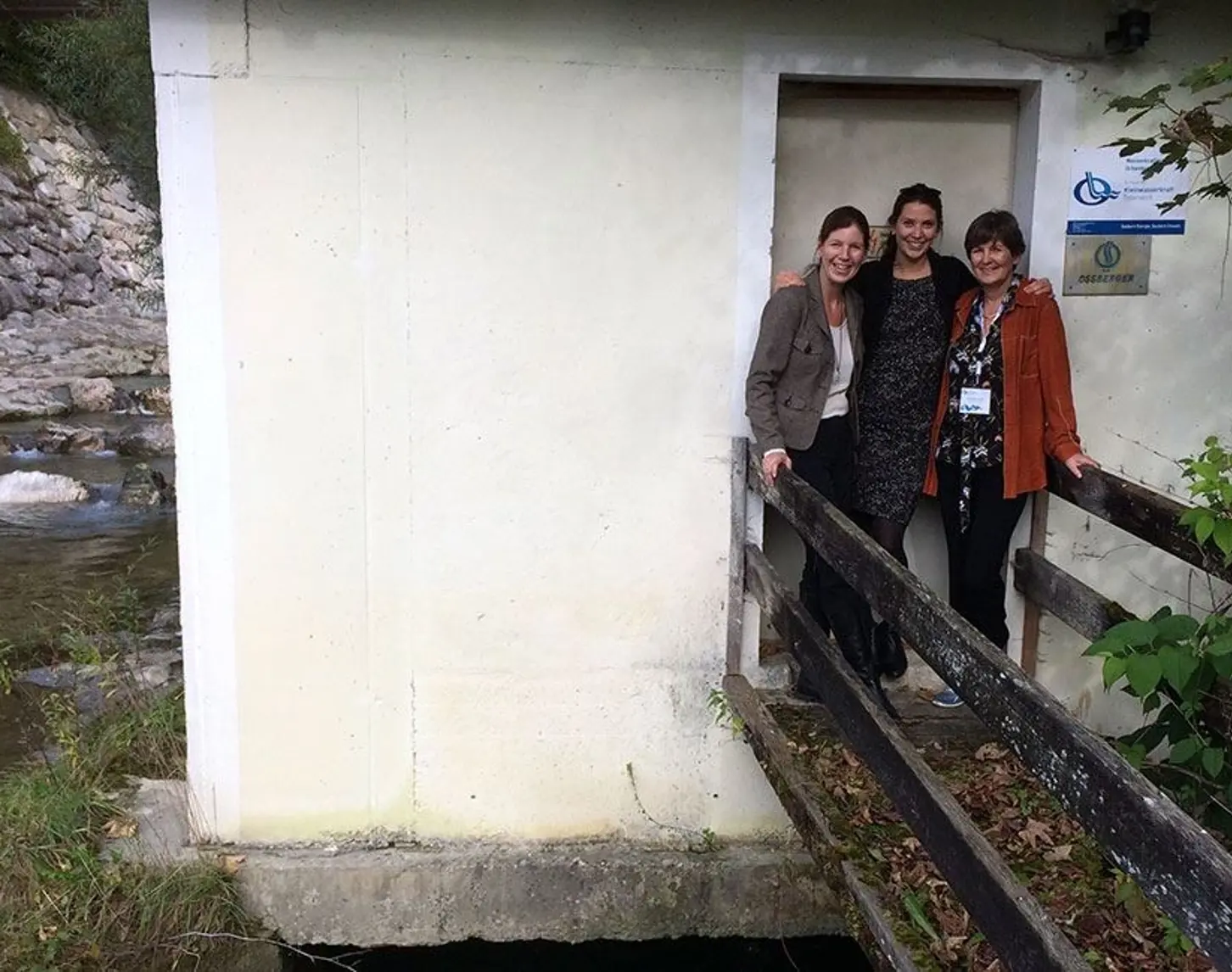 Drei Frauen stehen vor dem Eingang zu einem kleinen gelben Haus und lächeln.