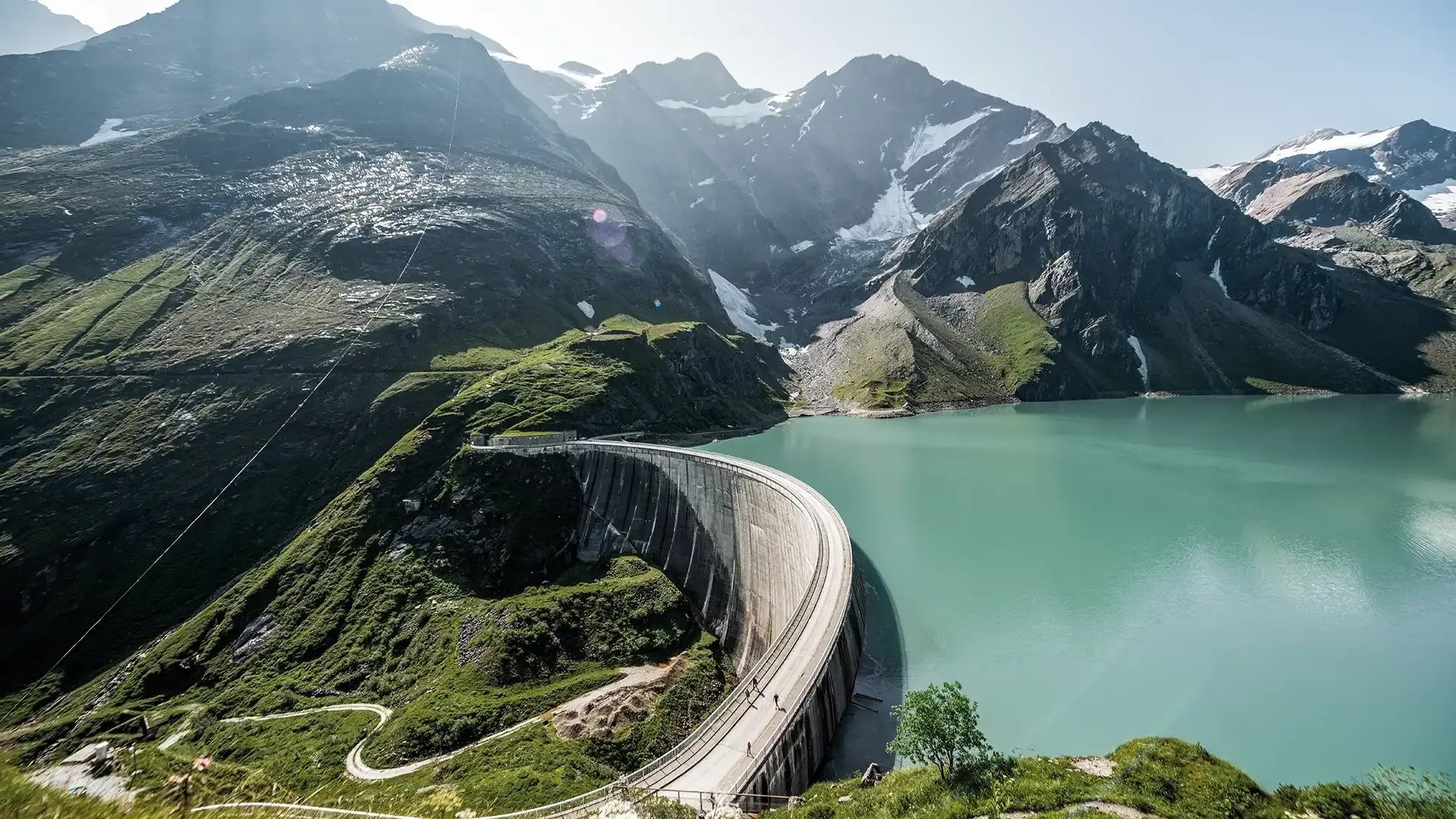 An aerial view of a dam in an alpine landscape, the storage lake is of a clear turqoise.