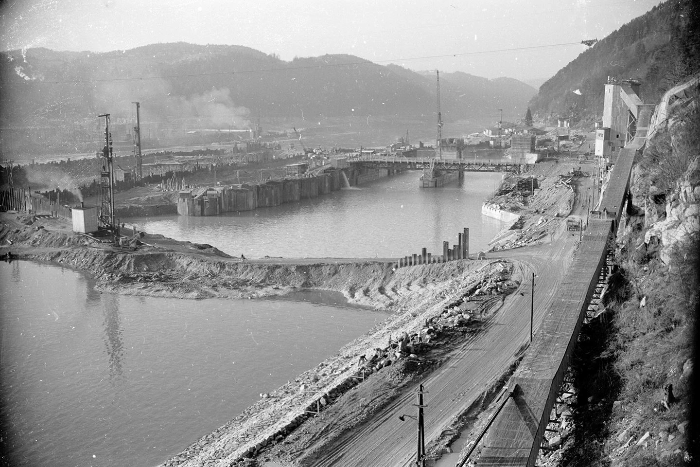 This is a historical photograph of the construction of the Aschach power station in black and white. Scaffolding, piles of gravel and cranes can be seen both in and next to the river. To the right of the river is a temporary shaft