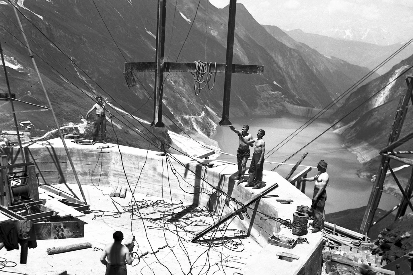 This is a historical photo of workers building the Kaprun power station.  Heavy steel scaffolding is lifted onto the dam walls with a crane.