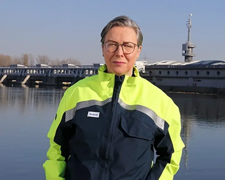 Dorothea Misch-Balanjuk steht mit einer Schutzjacke in grellem Gelb und dunkelblau mit silbernen Reflektorstreifen vor einem Wasserkraftwerk bei Sonnenschein.