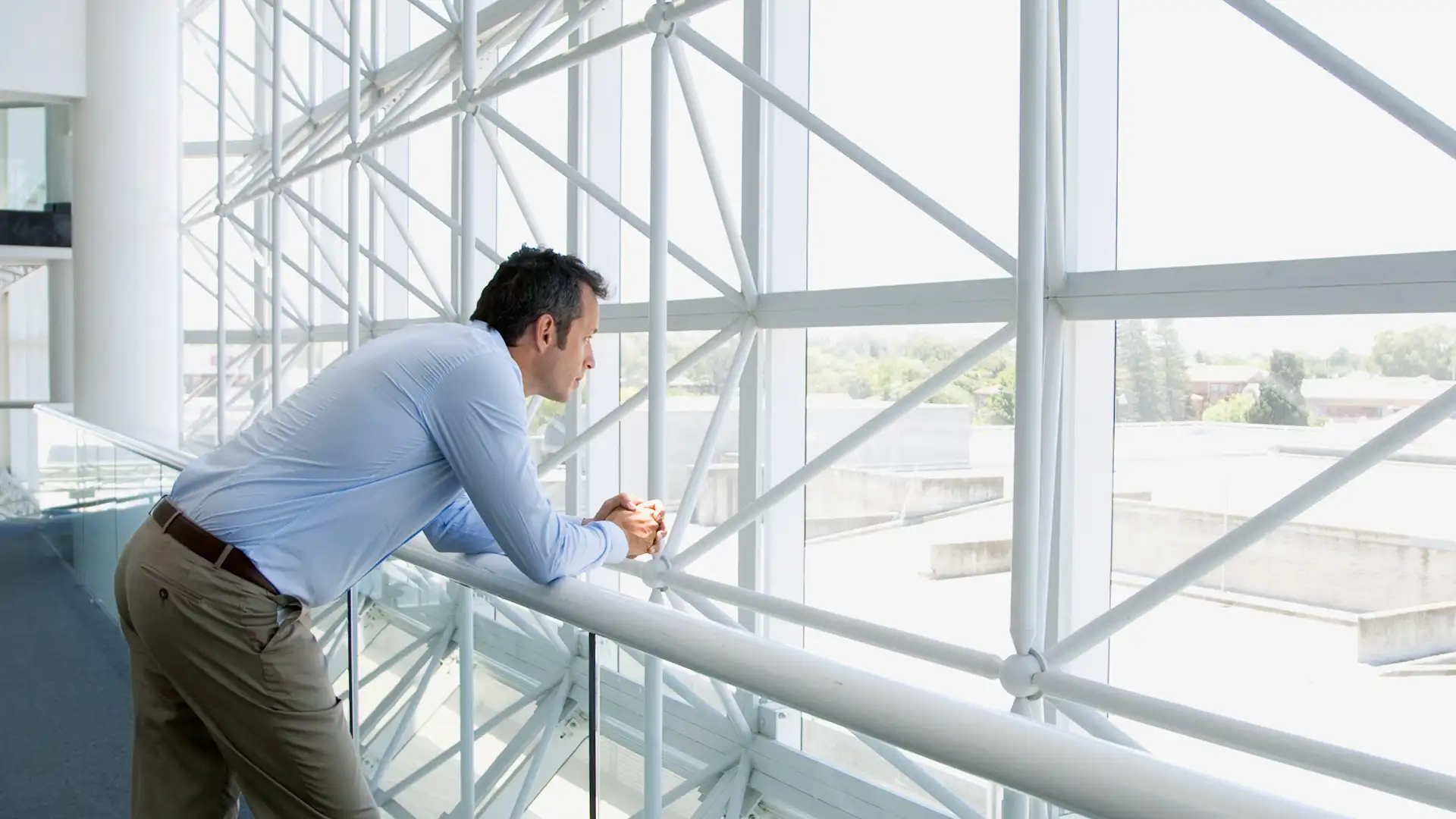 Businessman thoughtfully looking out of a large window in a modern office building. He has his arms resting on the railing and appears contemplative.