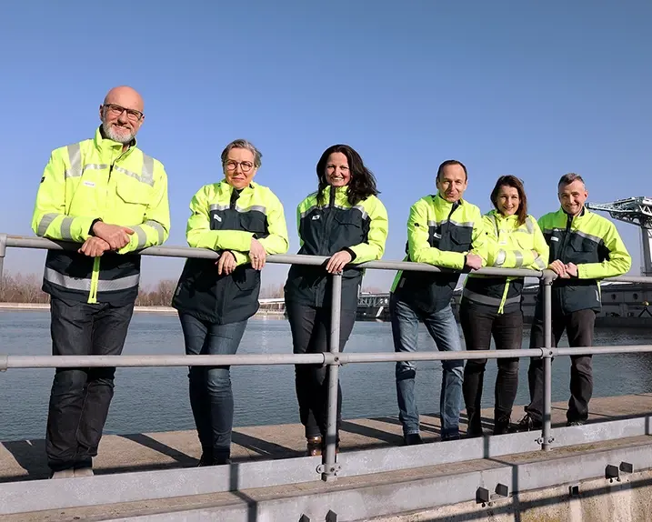 The PR team is standing on a bridge near a hydroelectric power station. They are standing in a row wearing safety jackets in dark blue and neon yellow. Right at the front is Rainer Tschopp. He wears glasses and a white beard. Next to him is Doro Misch-Balanjuk. Her hair is light-coloured and she also wears glasses. To her right is Ingun Metelko, she is smiling and has long brown hair. Robert Zechner is leaning against the railing next to her. His hair is cut short and dark brown. To his right is Eveline Fitzinger with medium-length brown hair. Next to her is Florian Seidl with medium brown hair. He is also smiling.