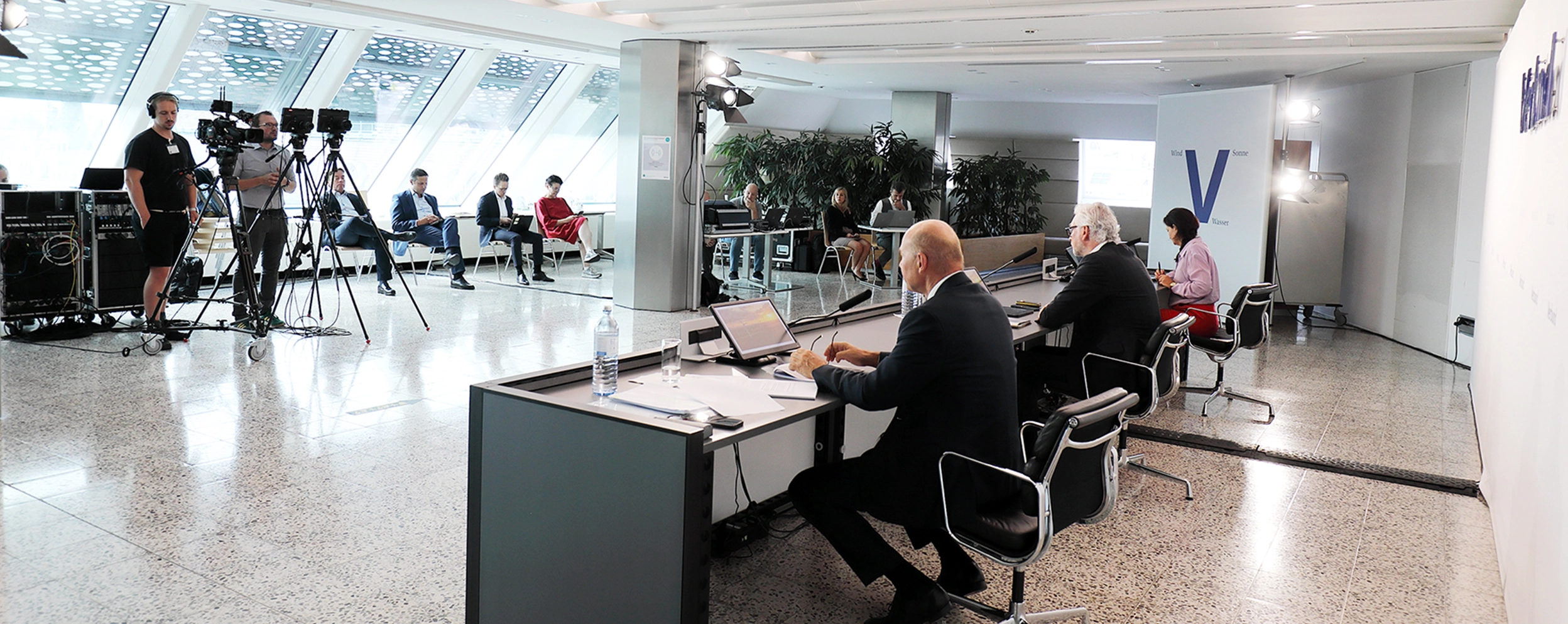Wir blicken von hinten auf eine Pressekonferenz mit Herrn Kollmann, Herrn Stugl und Frau Metelko. Wir sehen sie etwas seitlich. Am rechten Bildrand sind Journalisten und ein kleines Kamerateam zu sehen.