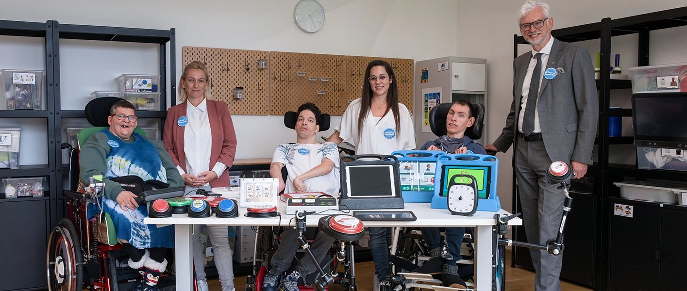 Auf dem Bild sind Mitarbeiter und Mitarbeiterinnen der Diakonie zu sehen in einer Werkstatt. Ganz links eine älter Dame mit grauen kurzen Haaren und blauem Kleid im Rollstuhl, neben Ihr eine Dame mit blonden Haaren und weißer Bluse mit rosa Jacket, etwas mittiger sitzt ein Mann mit weißem Oberteil und dunklen kurzen Haaren,  rechts von ihm eine Dame in weißem Oberteil mit braunen langen Haaren und Brille, rechts von ihr ein junger Mann mit dunklem Pullover, der zwei Bildschirme hält. Ganz rechts steht VERBUND CEO Michael Strugl, mit grauem Anzug, weißem Hemd und dunkelgrauer Kravatte. Er trägt eine Brille und hat weißes Haar.