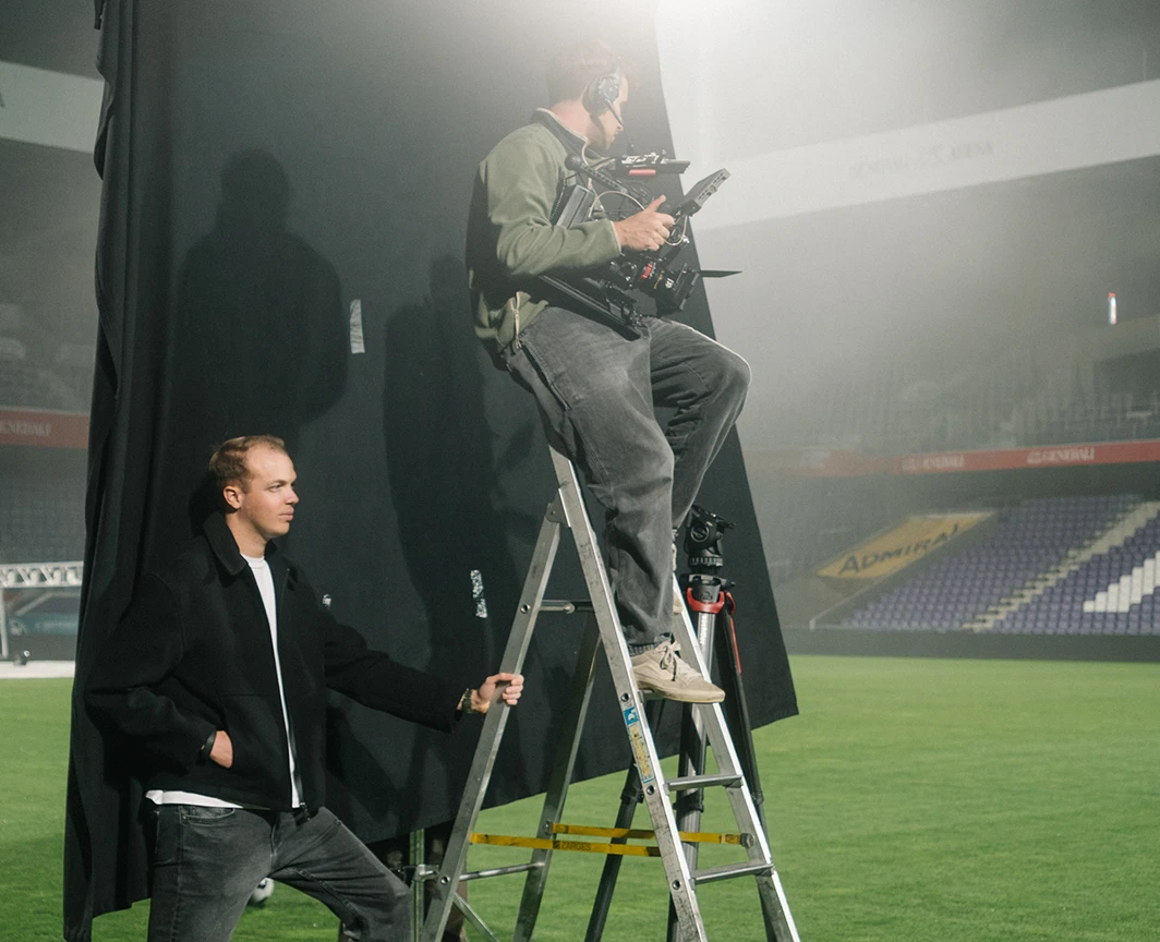 Zwei Männer arbeiten auf einem Fußballfeld mit Kamera- und Lichttechnik; einer steht auf einer Leiter, der andere hält einen schwarzen Stoff hinter ihnen.