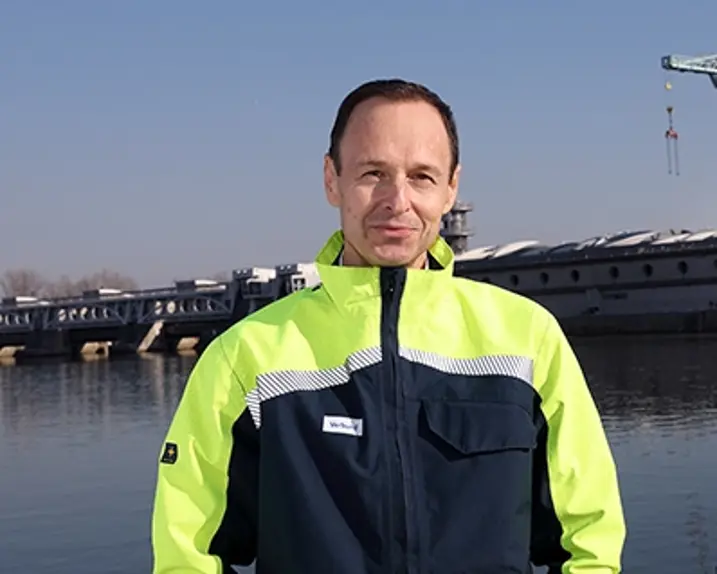 Robert Zechner steht vor einem Wasserkraftwerk. Er trägt ein Lächeln und eine Schutzkleidung in grellem gelb und dunkelblau mit silbernem Reflektorstreifen.