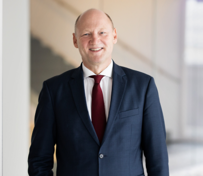 Achim Kaspar lächelt in die Kamera. Er trägt ein weißes Hemd, eine bordeau-farbene Krawatte und einen dunkelblauen Anzug.