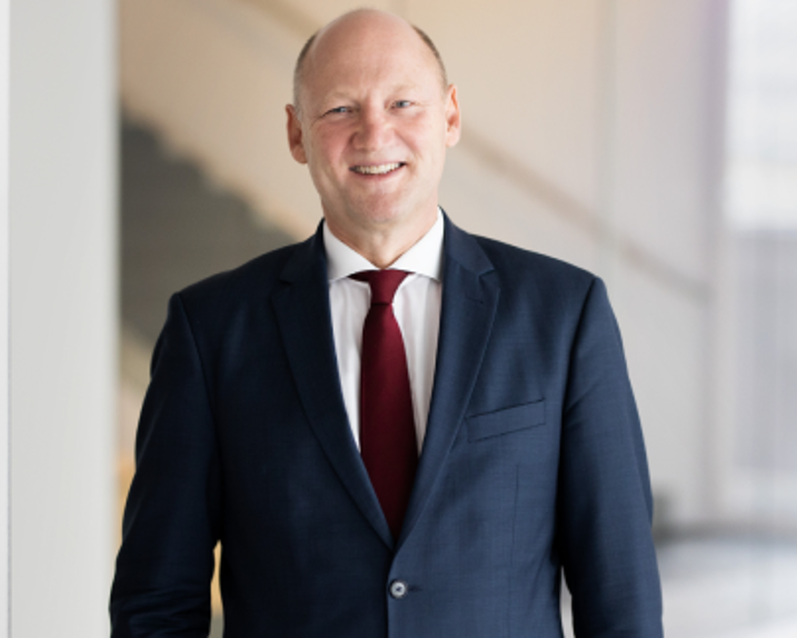 Achim Kaspar lächelt in die Kamera. Er trägt ein weißes Hemd, eine bordeau-farbene Krawatte und einen dunkelblauen Anzug.