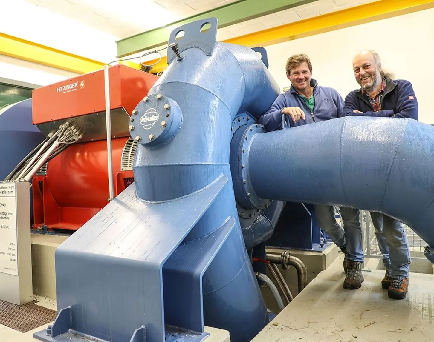 Zwei Männer, die hinter einer Turbine in einem Wasserkraftwerk stehen.