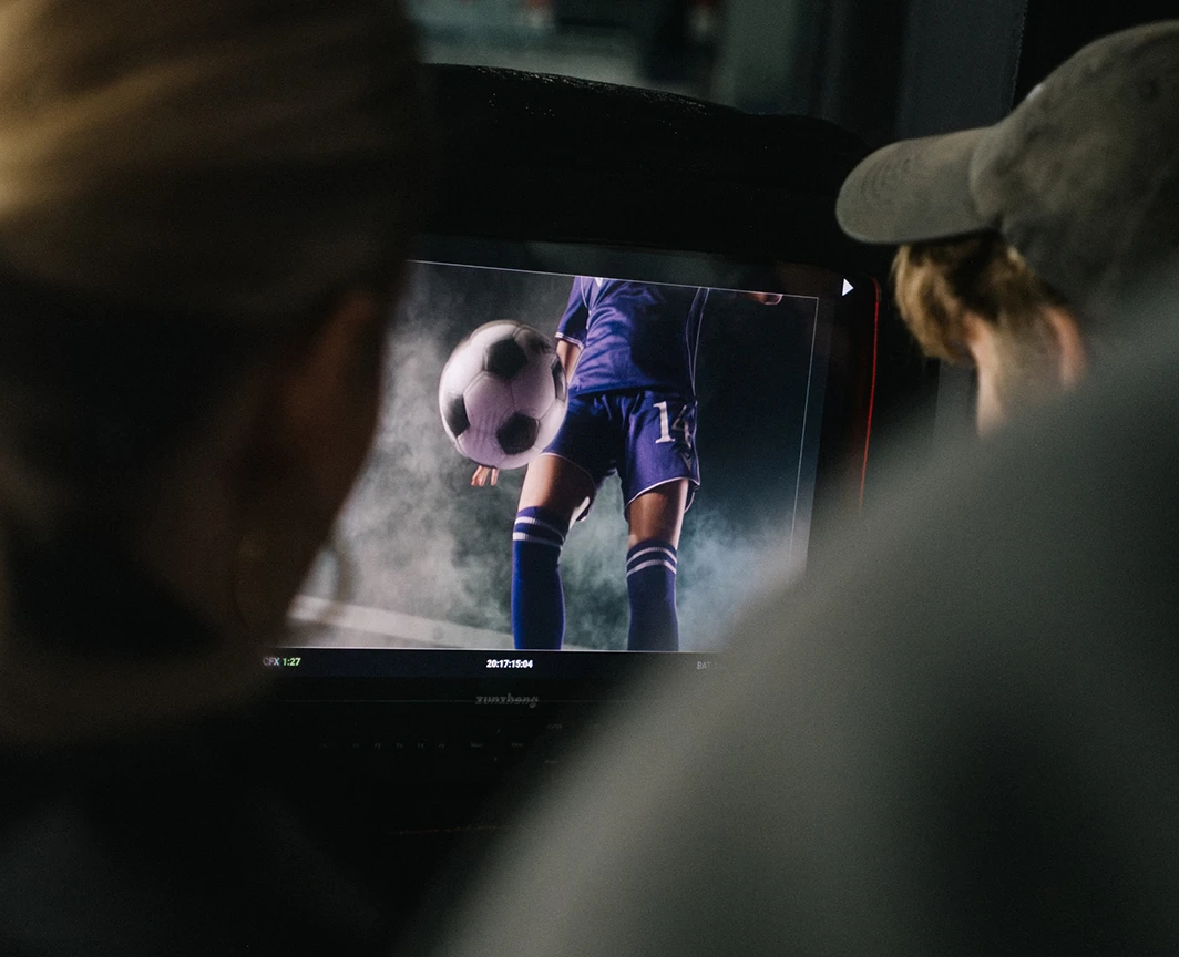 Zwei Personen sehen auf einen Monitor mit einer Fußballspielerin im violeten Trikot und einem Ball in einer dunklen Szene.