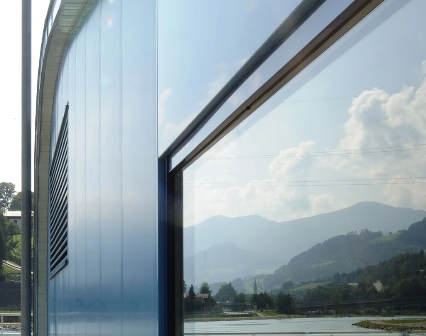 We see the window front of an office building to the right of the picture. The windows reflect nature with an electricity pylon above the woods. A lake lies in front of the office building.