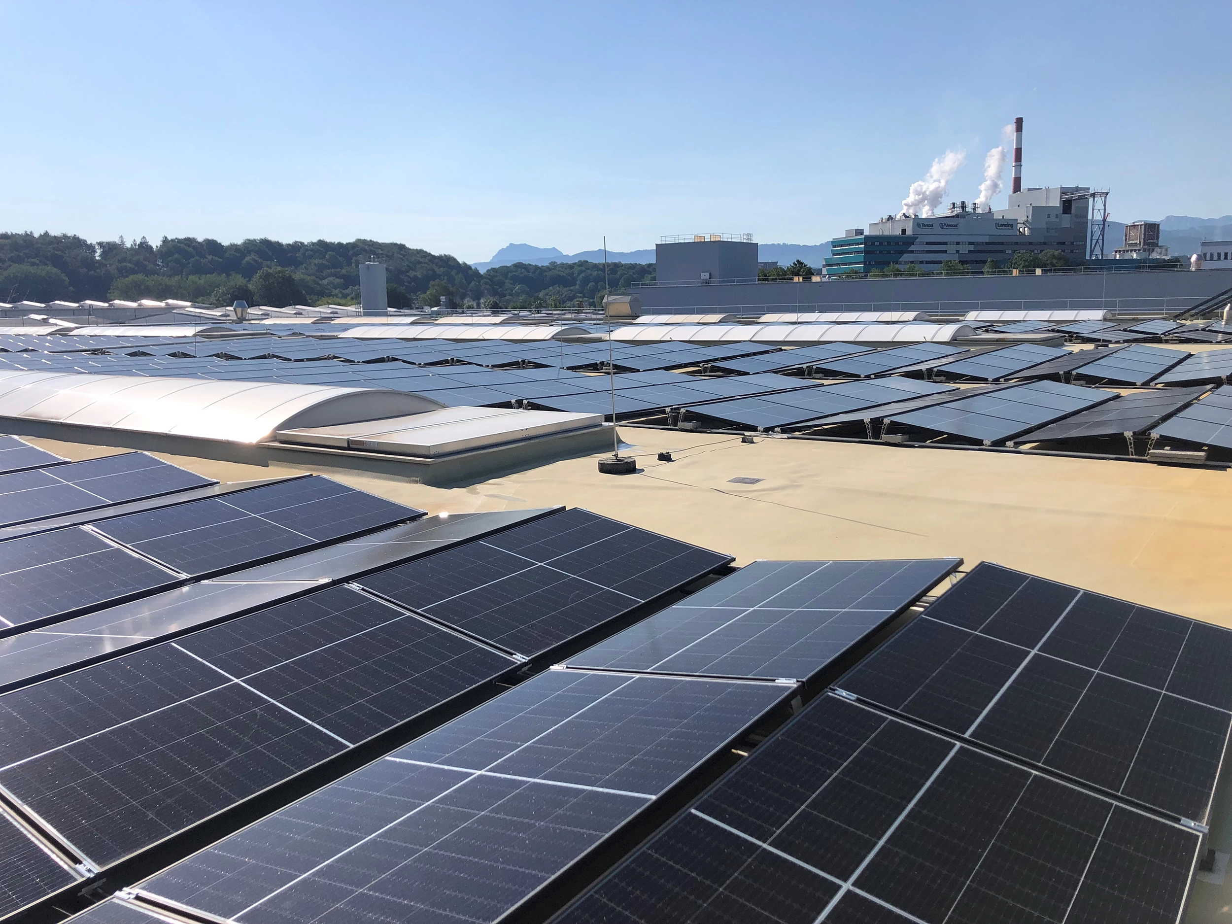 Large-scale photovoltaic system for industry on the roof of a hall. In the background you can see a factory with a steam vent. The sky is blue.