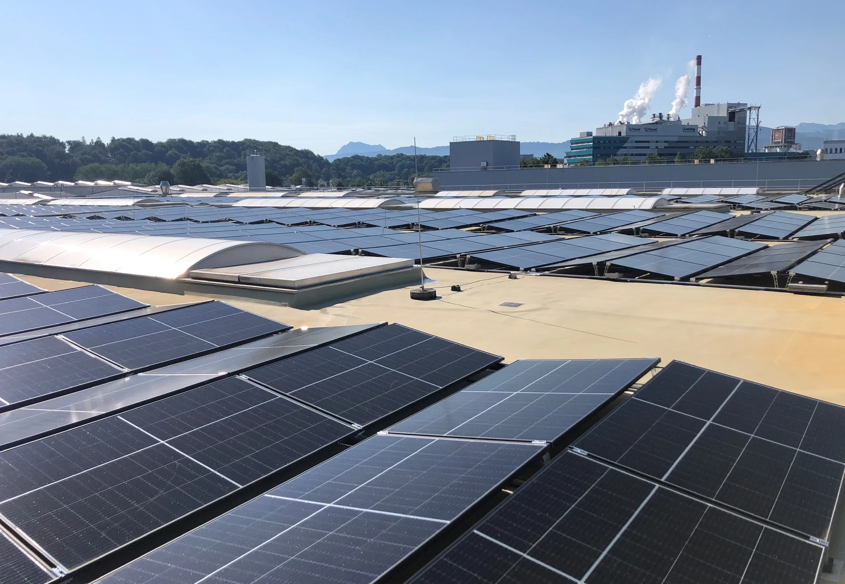 Photovoltaik Großanlage für die Industrie auch einem Hallendach. Im Hintergrund sieht man eine Fabrik mit Dampfschlot. Der Himmel ist blau.