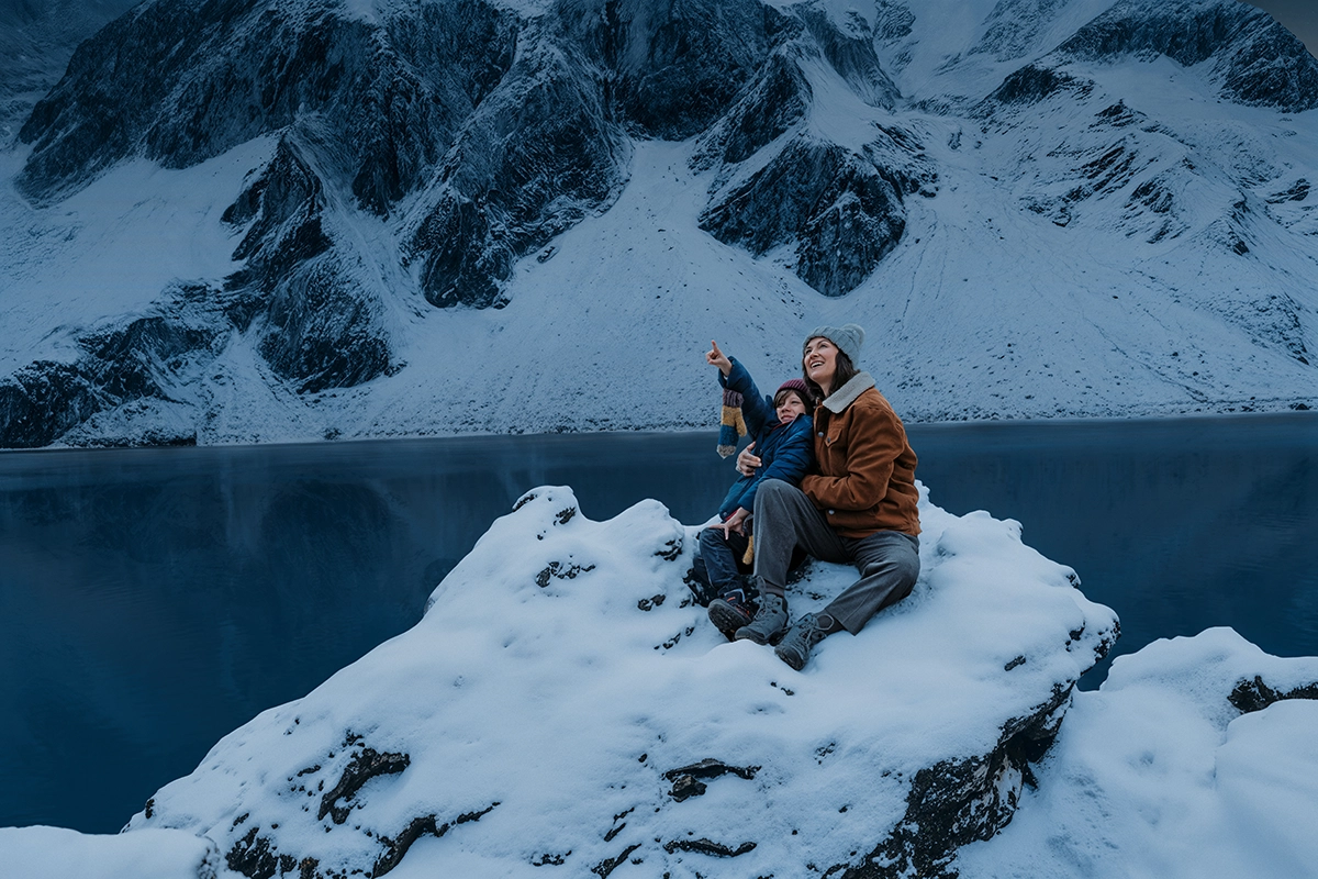 Wir blicken von oben auf Mutter uns Sohn. Beide tragen Winterklamotten und sitzen auf einem schneebedeckten Stein an einem Stausee. Sie lächeln und blicken in den Himmel während der Sohn nach oben zeigt.