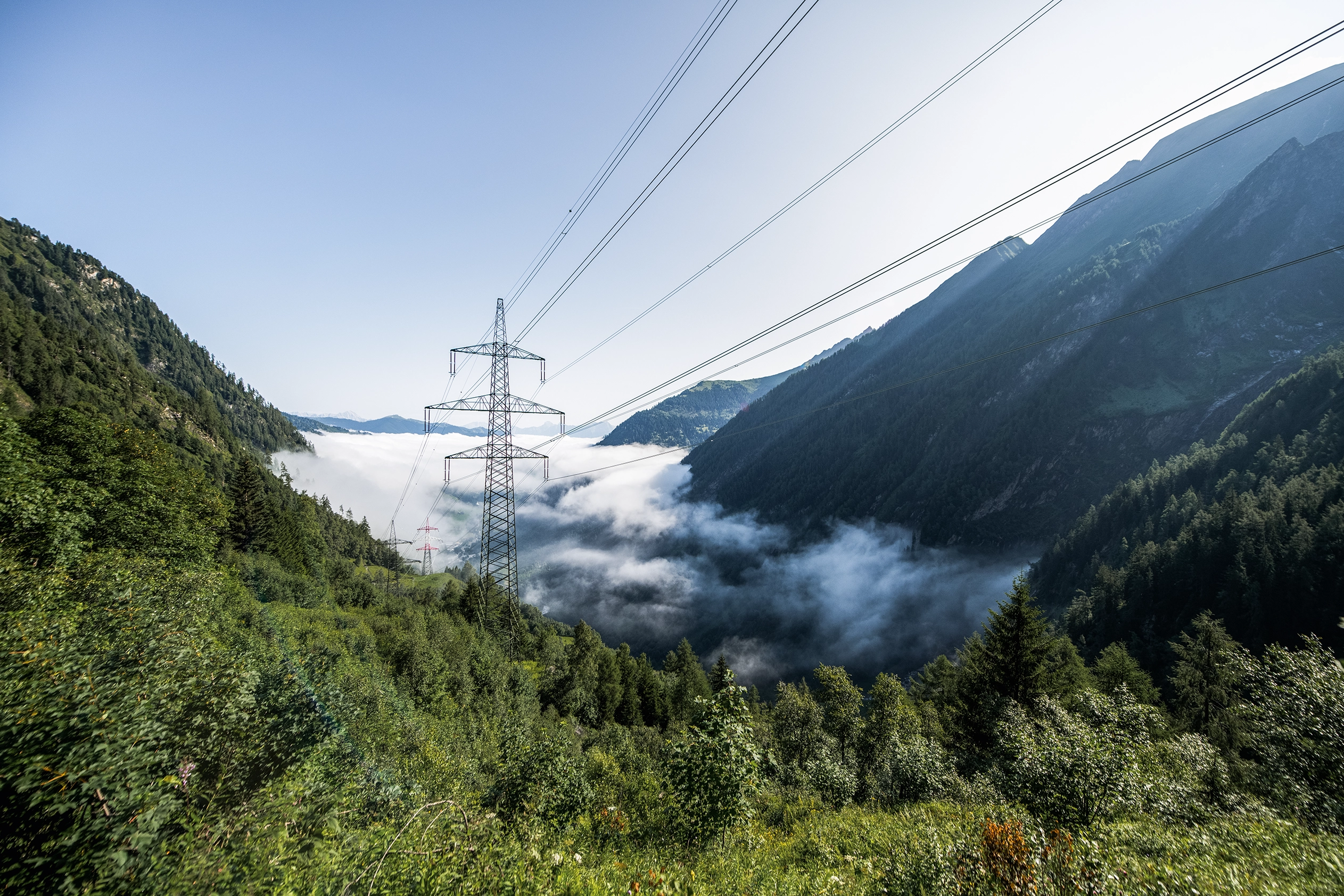 From above, we take a look down into a mist-covered valley that is lush and green. An electricity mast can be seen in the center.