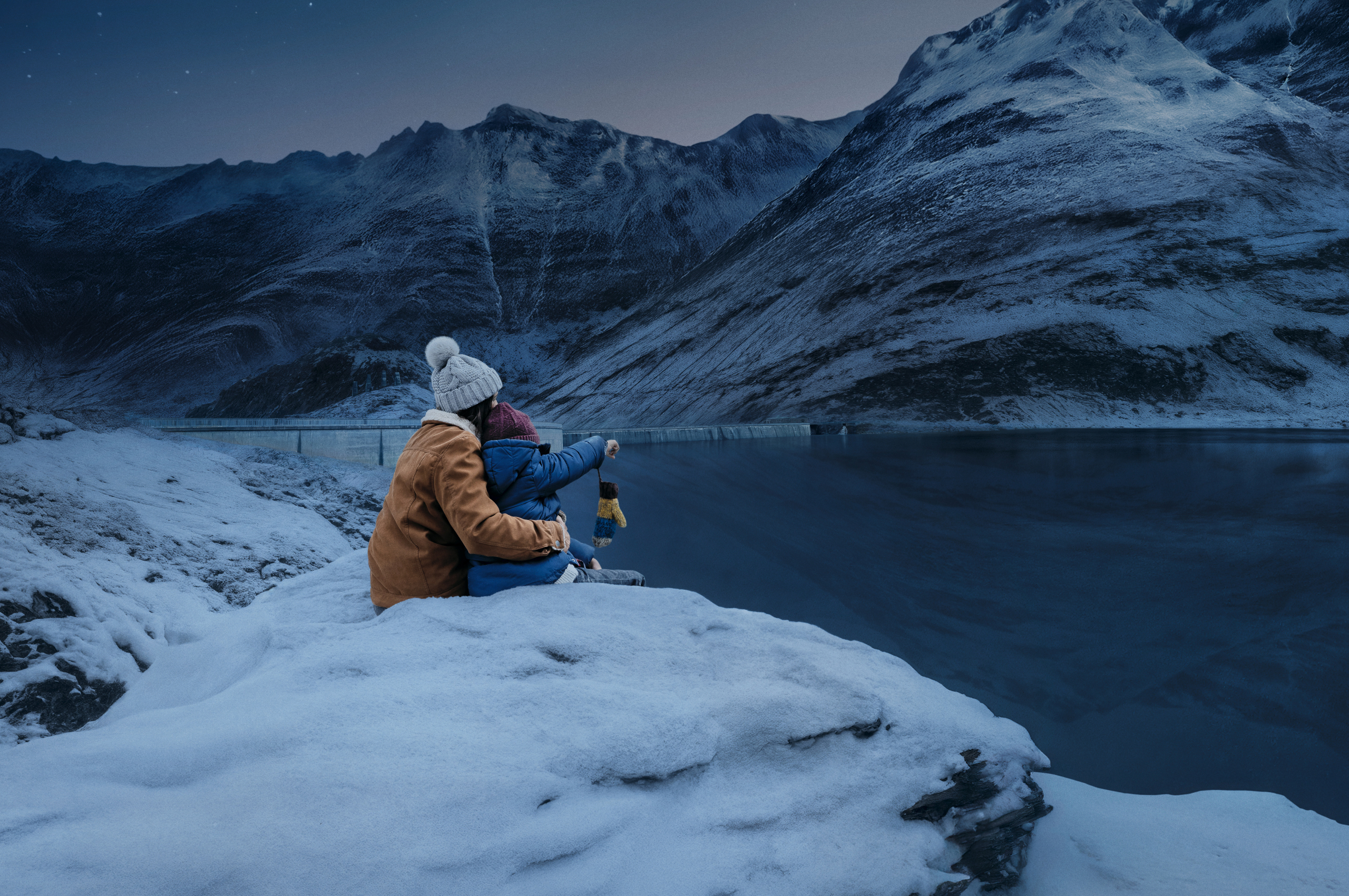 Eine Mutter mit hellgrauer Bommelmütze und brauner Winterjacke hält ihren Sohn. Er zeigt auf etwas in der Ferne. Wir sehen sie von hinten. Sie sitzen auf einem schneebedeckten Stein an einem Stausee.