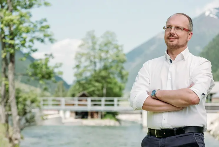 A person with crossed arms standing in front of a river and a bridge, surrounded by green trees and mountains.