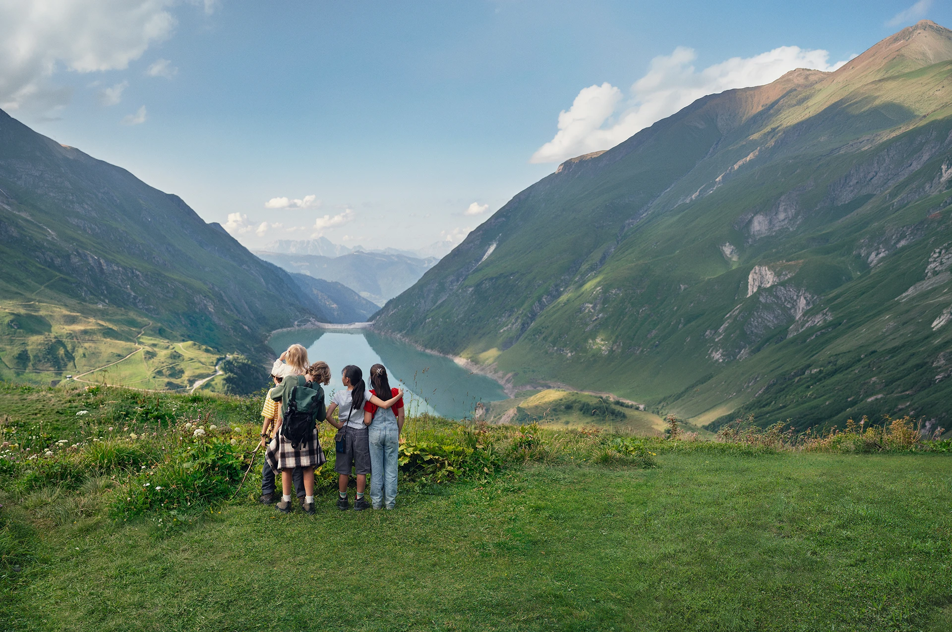 Five children stand on a grassy slope and look over an alpine valley with a lake between green mountains.