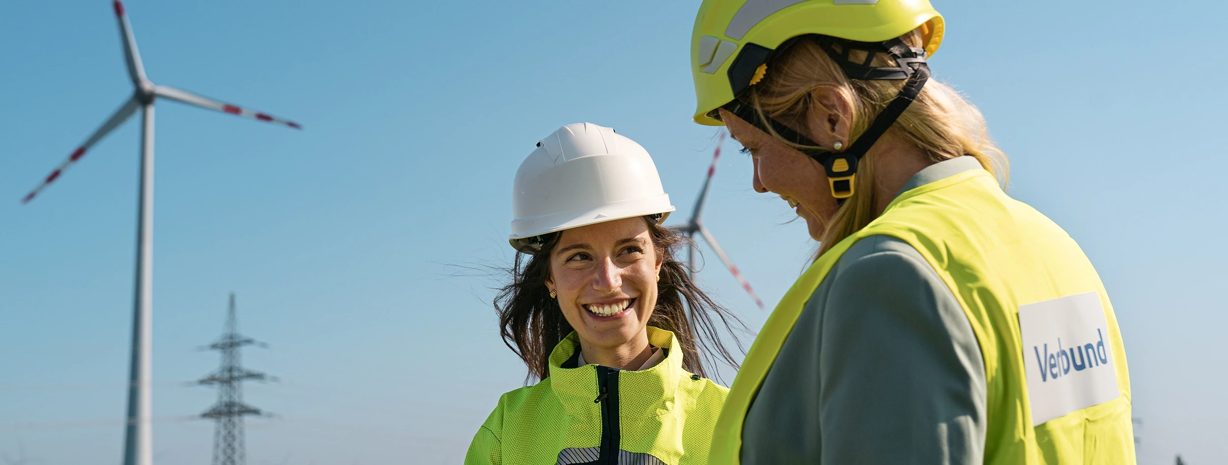Two women in safety clothing and helmets stand in front of two wind turbines, which can be seen slightly out of focus in the background. The sky is bright blue.