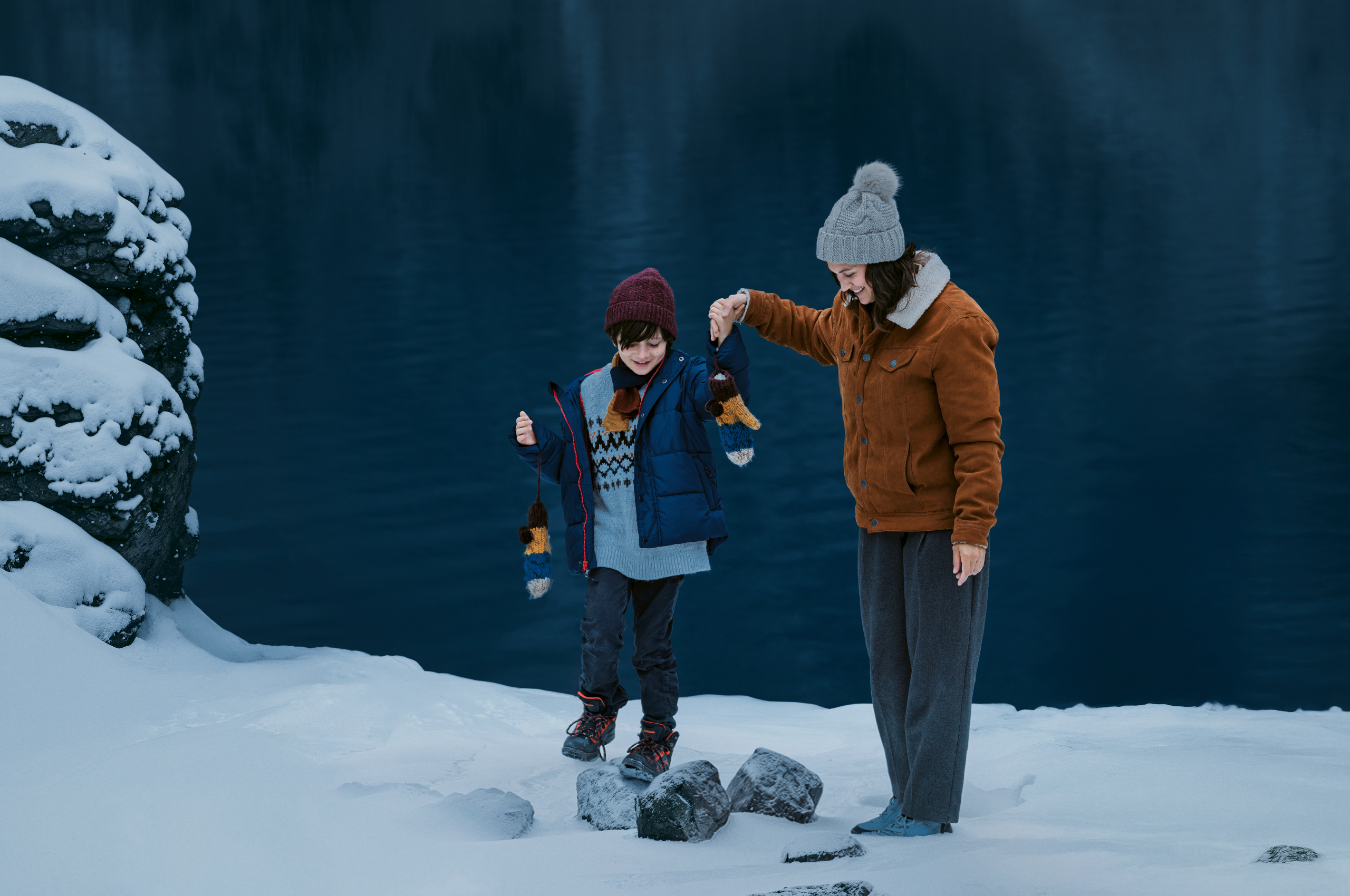 Mutter und Sohn spielen im Schnee an einem See. Beide tragen Mützen, Winterjacken und feste Schuhe. Die Sonne ist gerade untergegangen und es wird bereits Nacht.