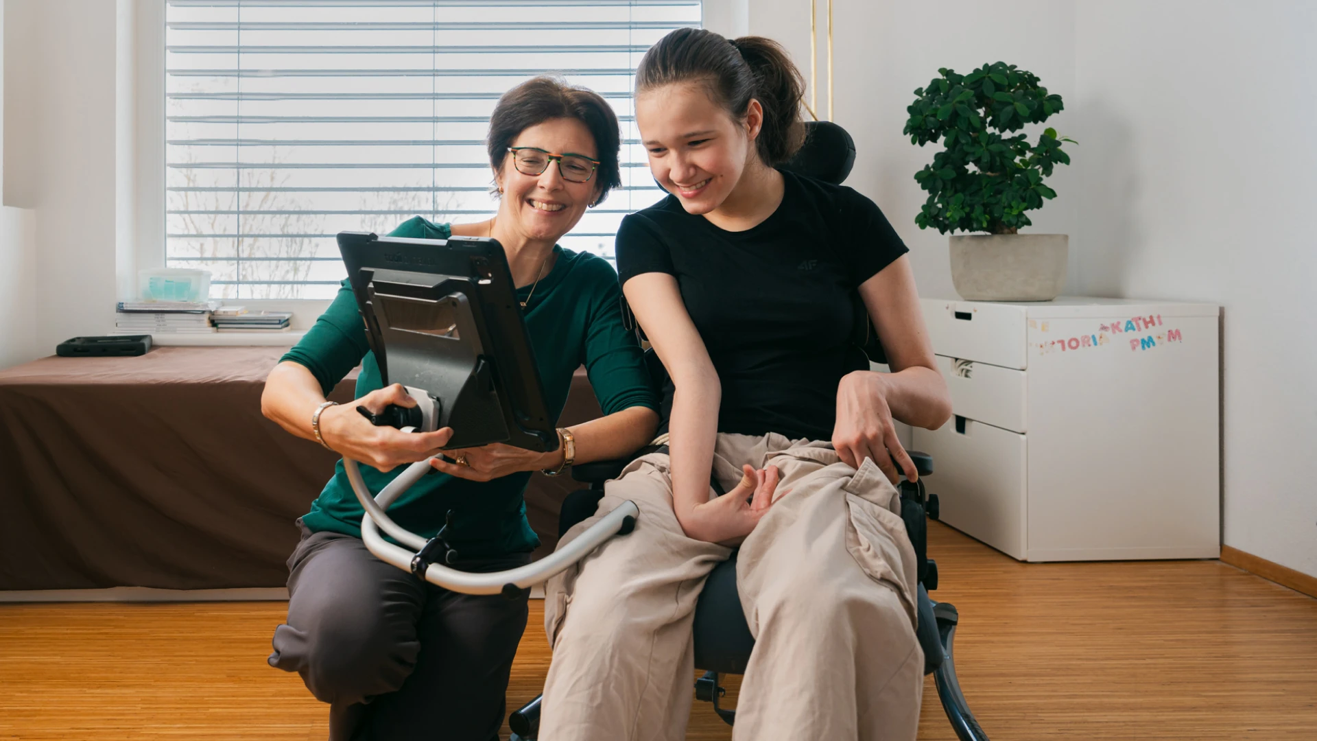 Mutter und Tochter blick auf einen Screen. Dieser ist direkt am Stuhl der Tochter angebracht. Beide lächeln.