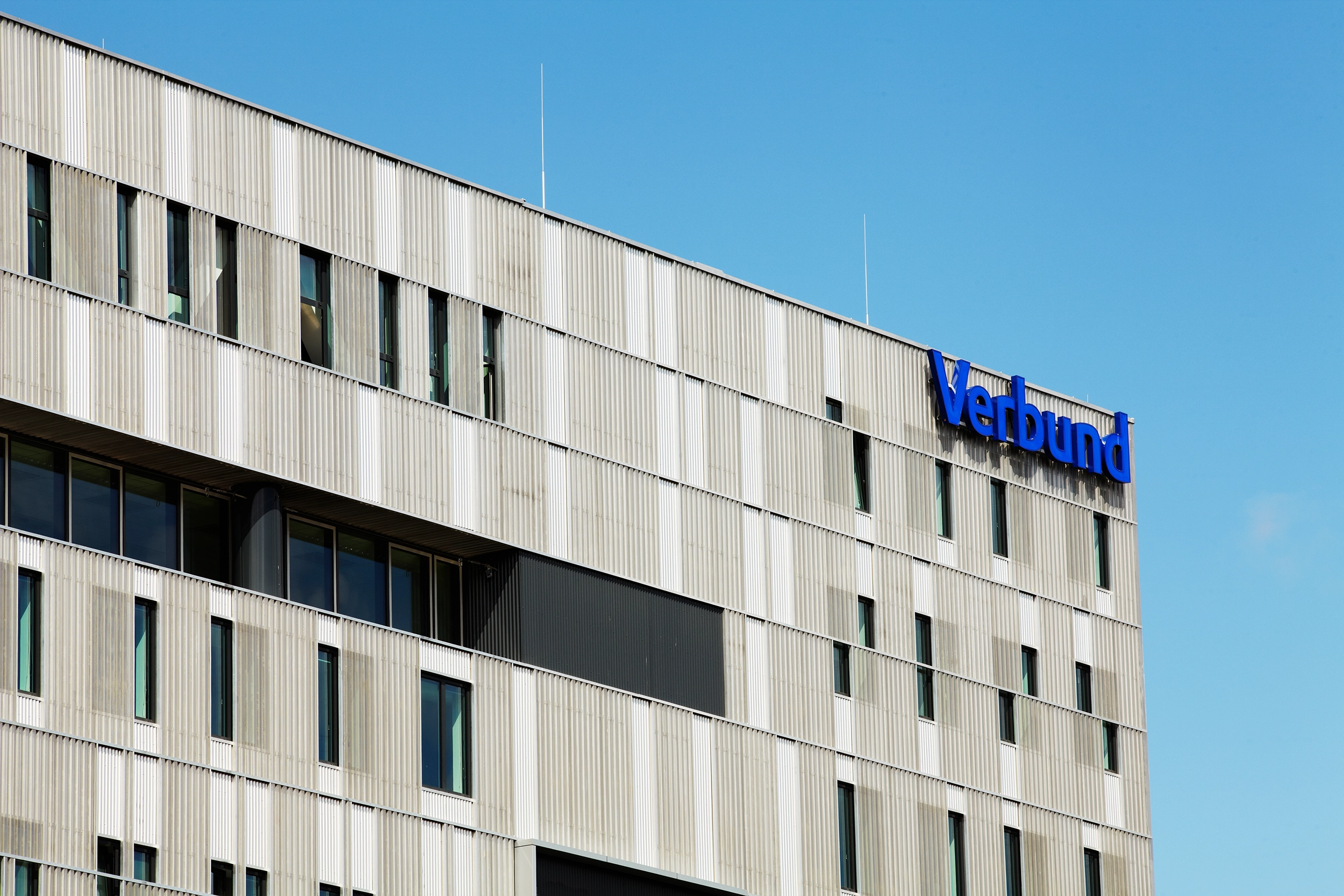 We look at the façade of VERBUND at Westbahnhof. It is grey with windows. At the top right is the VERSBUND lettering in VERBUND blue.