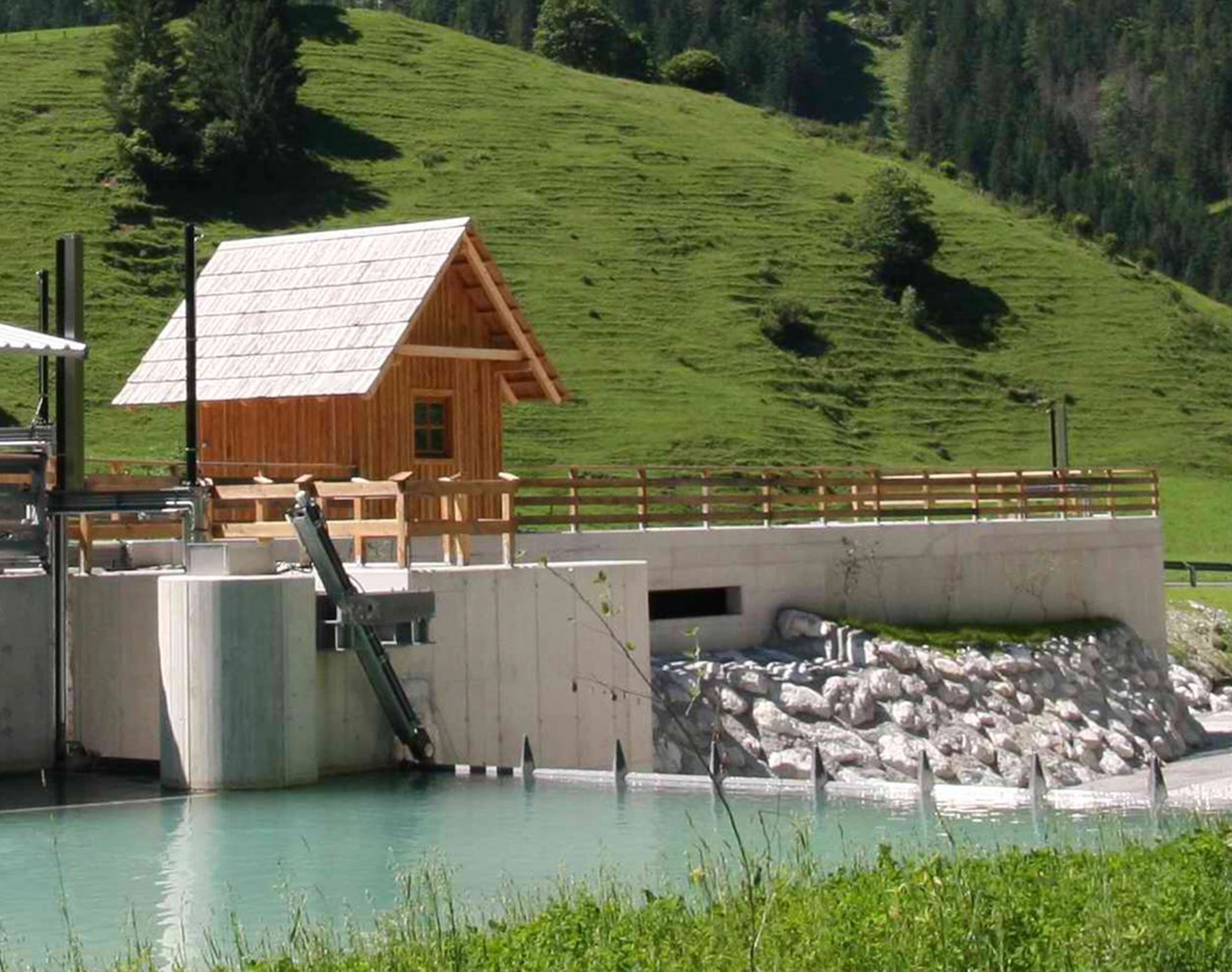 Wir blicken auf ein Kleinwasserkraftwerk mit saftig grünen Wiesen im Hintergrund.