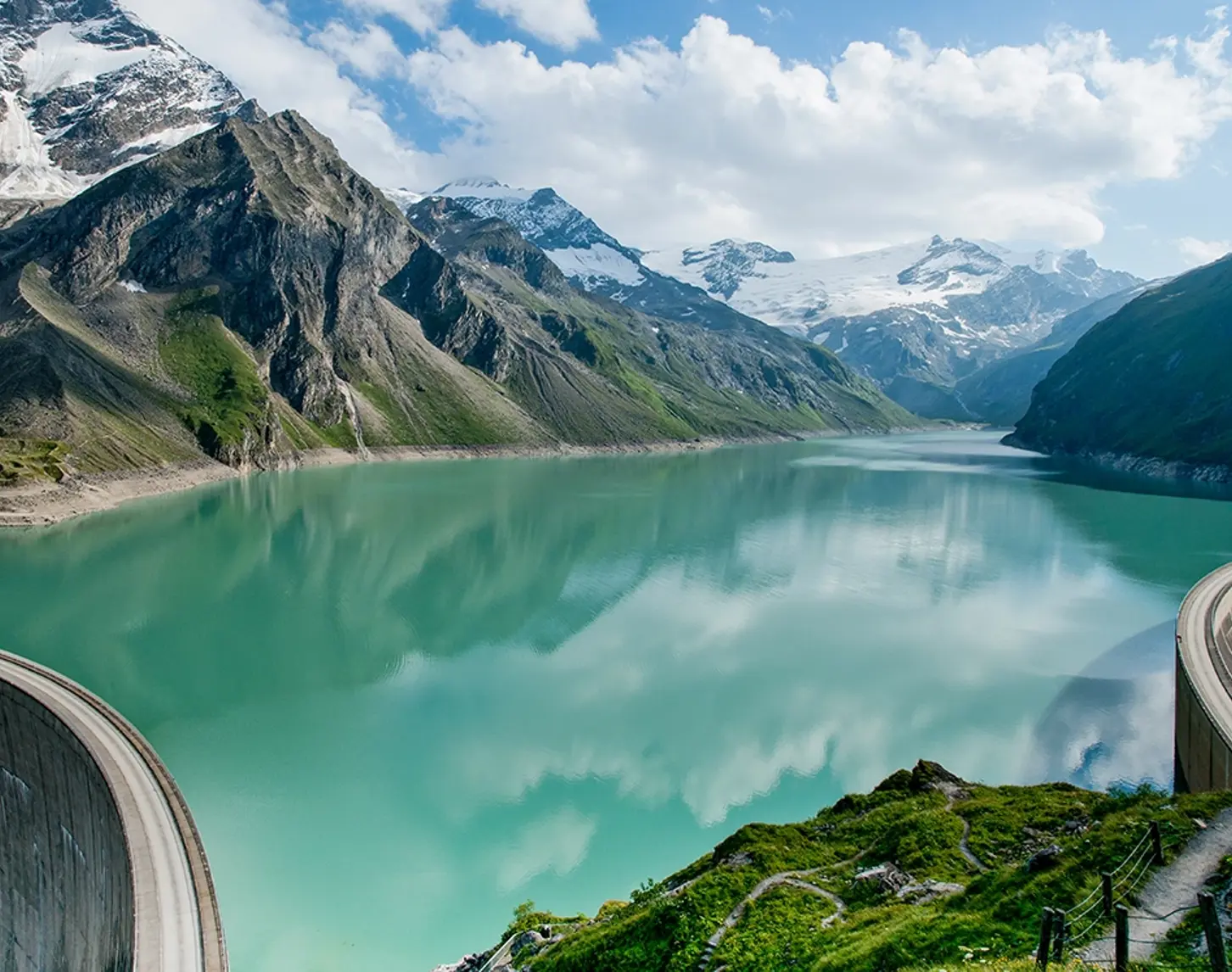 We look out over the Mooserboden reservoir in Kaprun. The clouds are reflected in the blue-turquoise water. The peaks in the background are covered in snow. The sun is shining and the meadows are green.