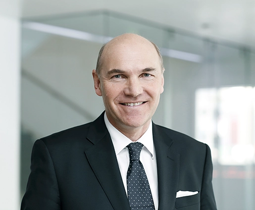 VERBUND-CFO Peter F. Kollmann trägt einen schwarzen Anzug und Krawatte. Er lächelt in die Kamera.