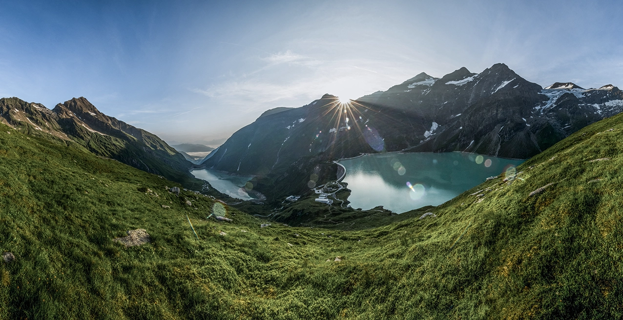 Wir blicken aus der Ferne auf den einen Sonnenaufgang im Kaprun. Die Wiesen sind herrlich grün. Der Stausee schimmert blau-türkis und die Sonne spitzt gerade mal über die Bergspitzen.