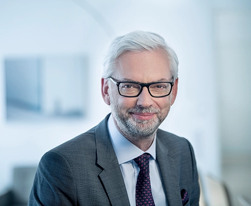 VERBUND-CEO Michael Strugl trägt einen grauen Anzug mit Krawatte. Er hat weißes Haar, eine Brille und einen graumelierten Bart. Er lächelt leicht.