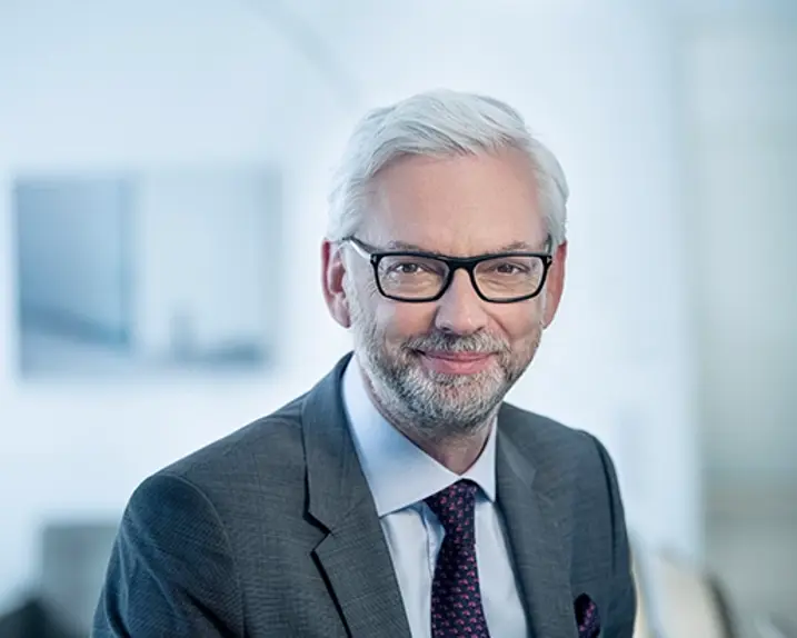 VERBUND-CEO Michael Strugl trägt einen grauen Anzug mit Krawatte. Er hat weißes Haar, eine Brille und einen graumelierten Bart. Er lächelt leicht.